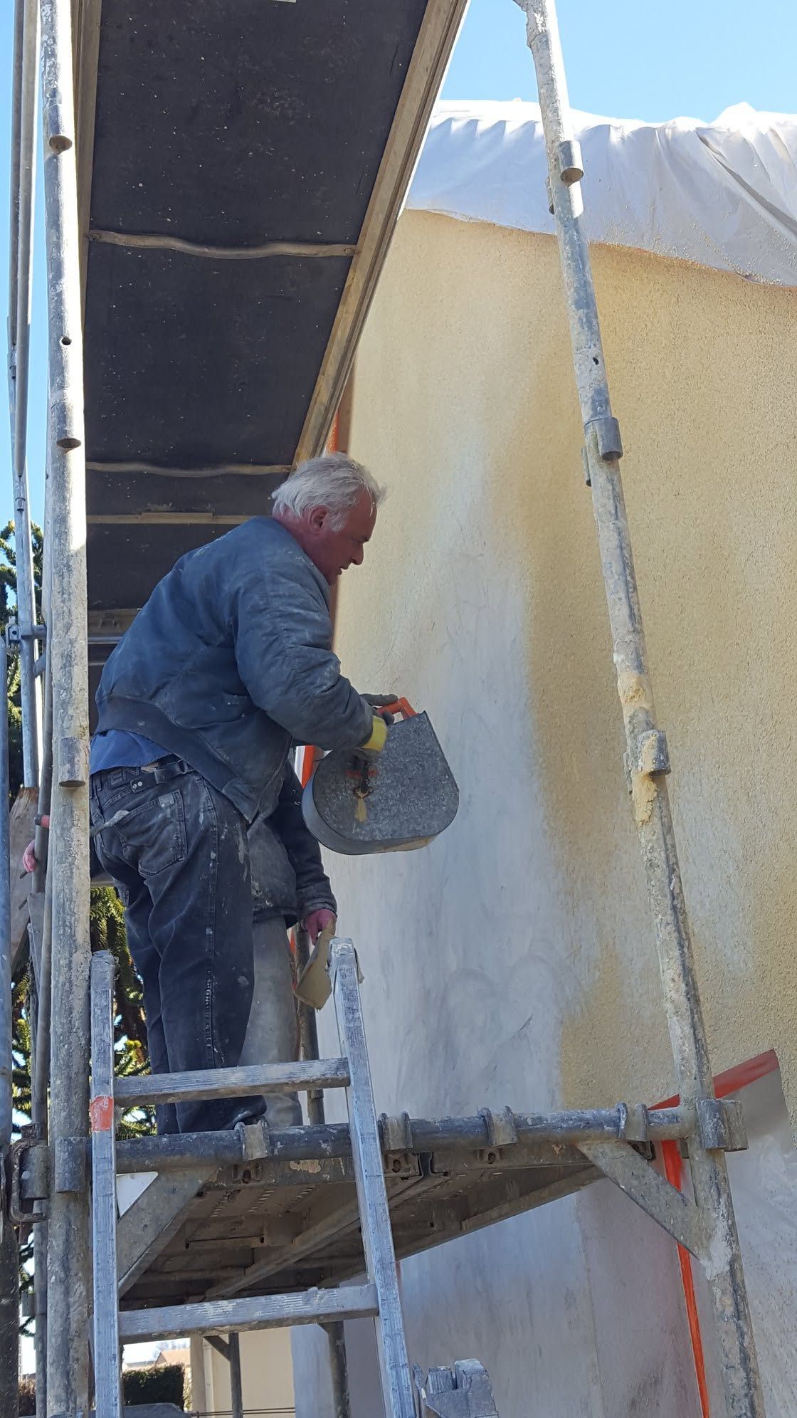 Ouvrier sur échafaudage appliquant du plâtre sur un mur sous une passerelle couverte.