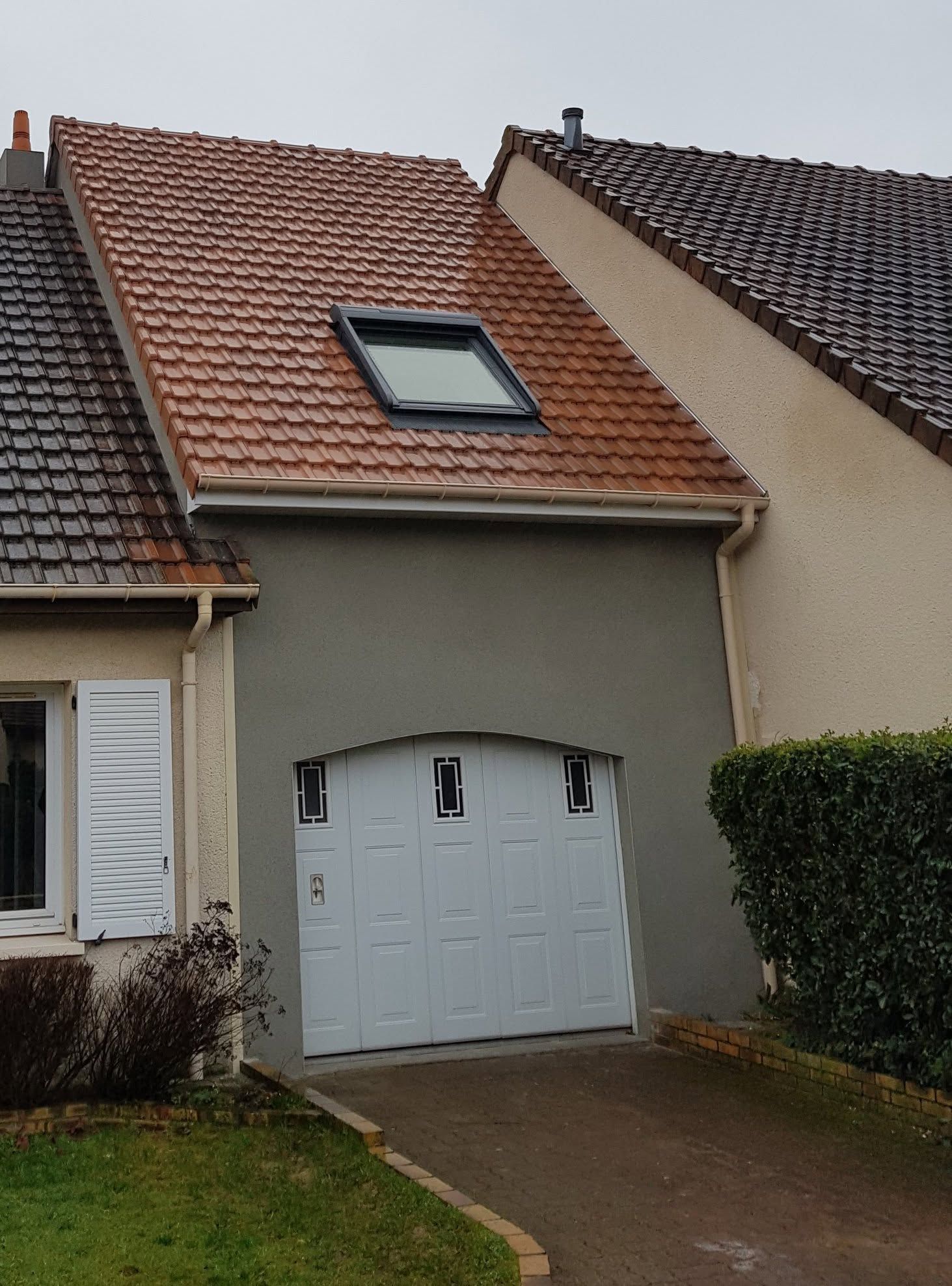 Extérieur de la maison avec une porte de garage blanche, un toit de tuiles rouges et un puits de lumière au-dessus de l'allée.