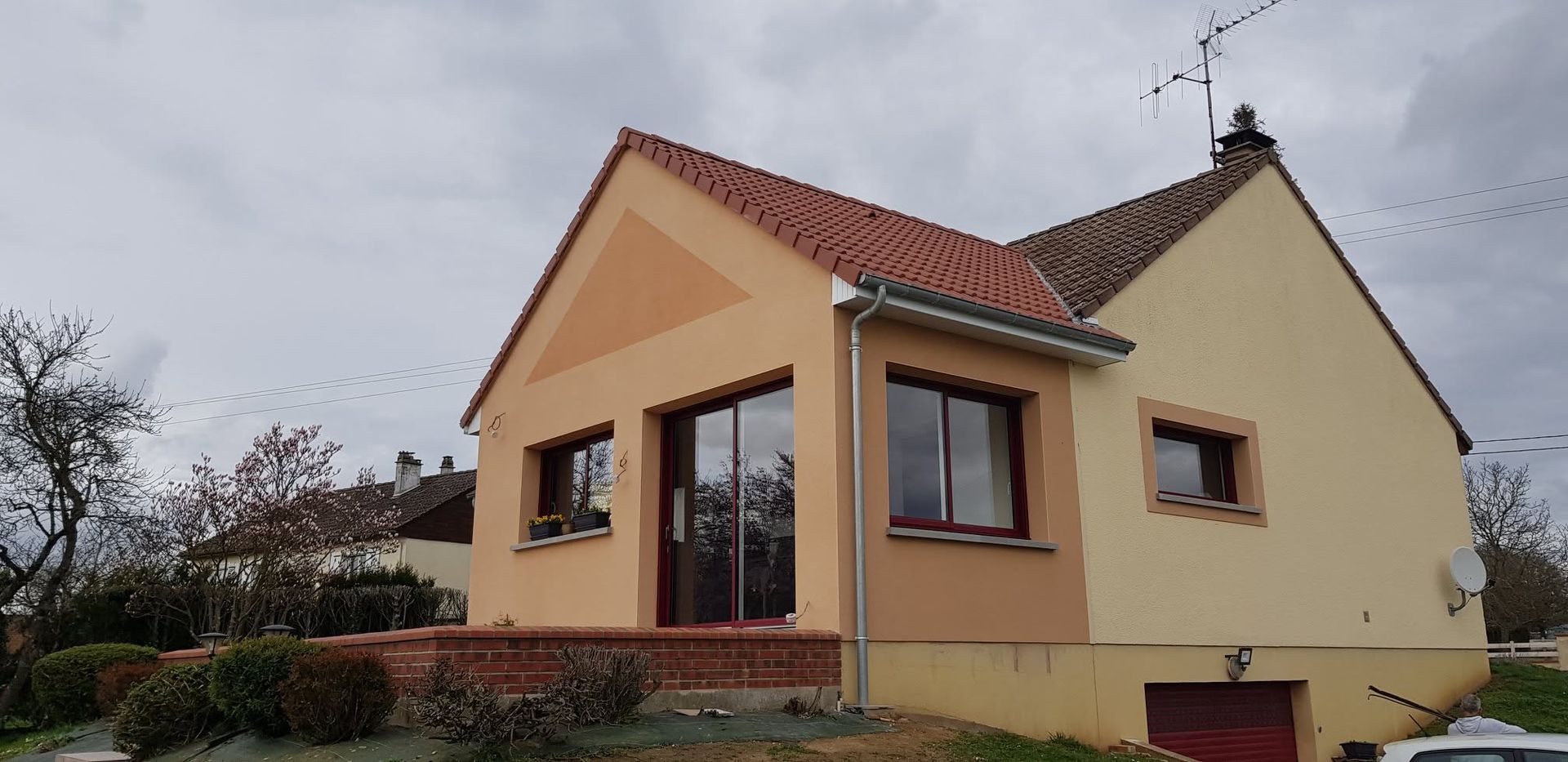 Petite maison beige au toit rouge et aux ornements en briques sous un ciel nuageux.