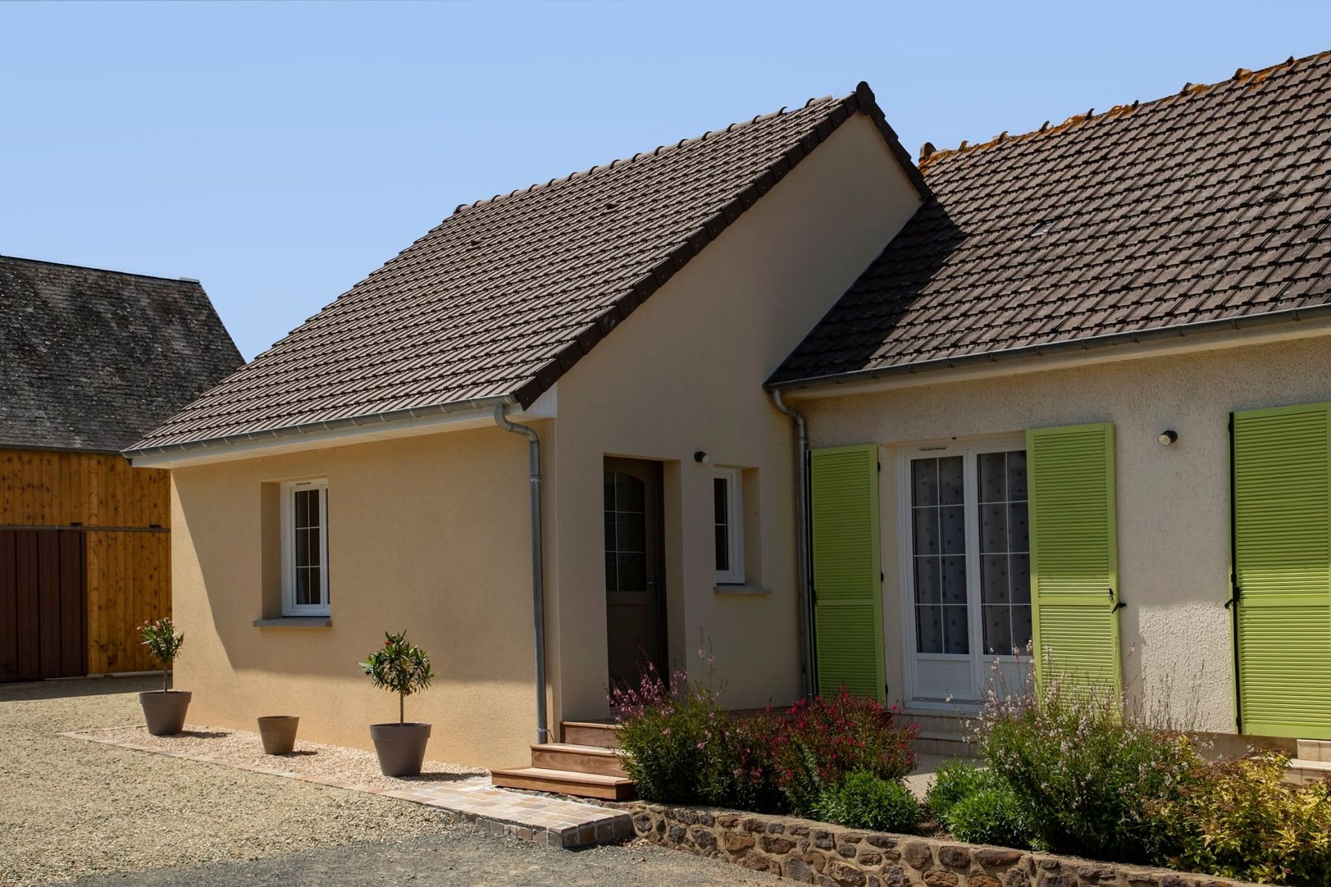Maison de plain-pied beige avec toit de tuiles brunes et volets verts dans une cour ensoleillée.