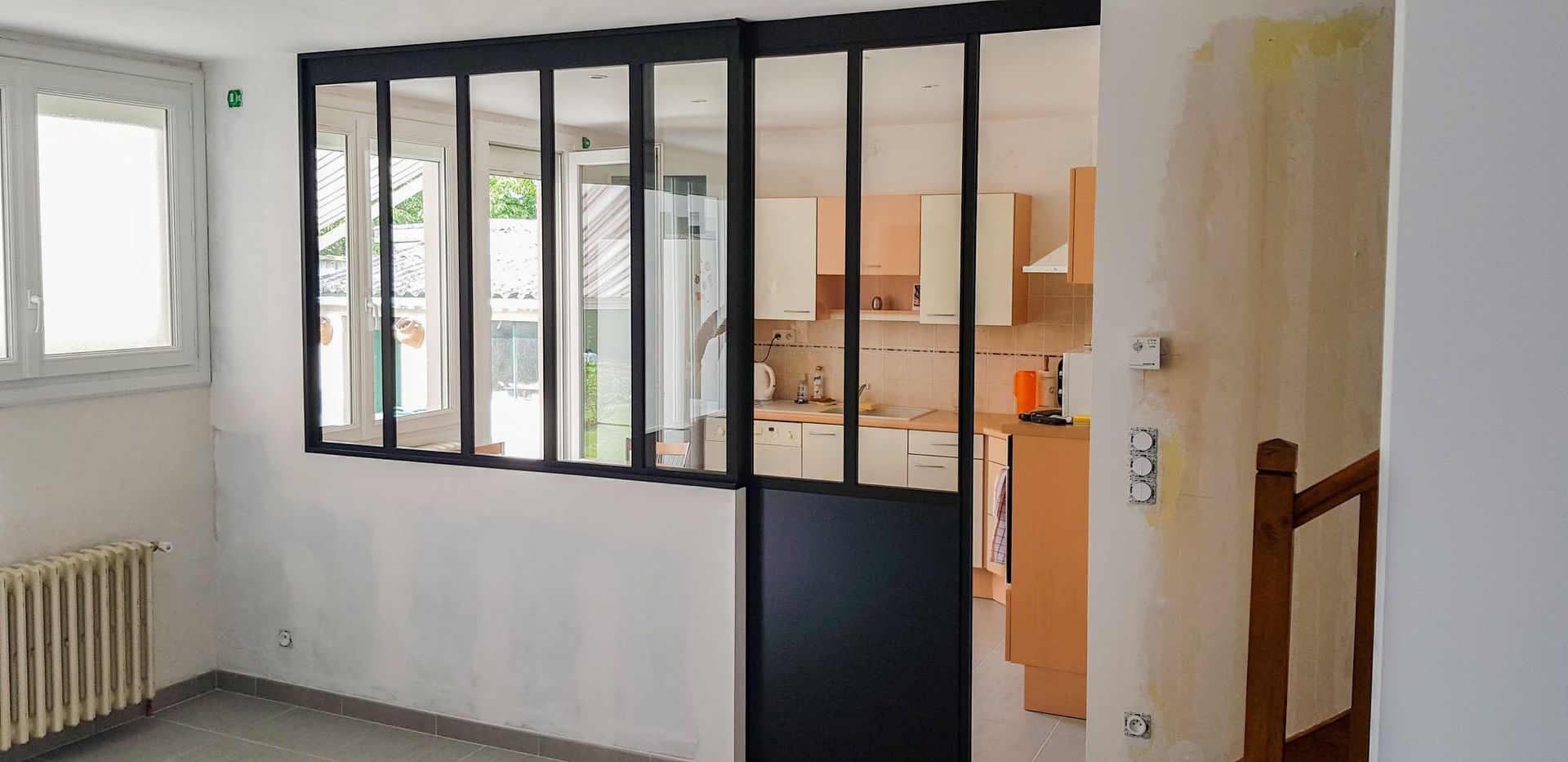 Pièce intérieure avec cloison vitrée à cadre noir, murs blancs, fenêtre, radiateur et porte donnant sur la cuisine.