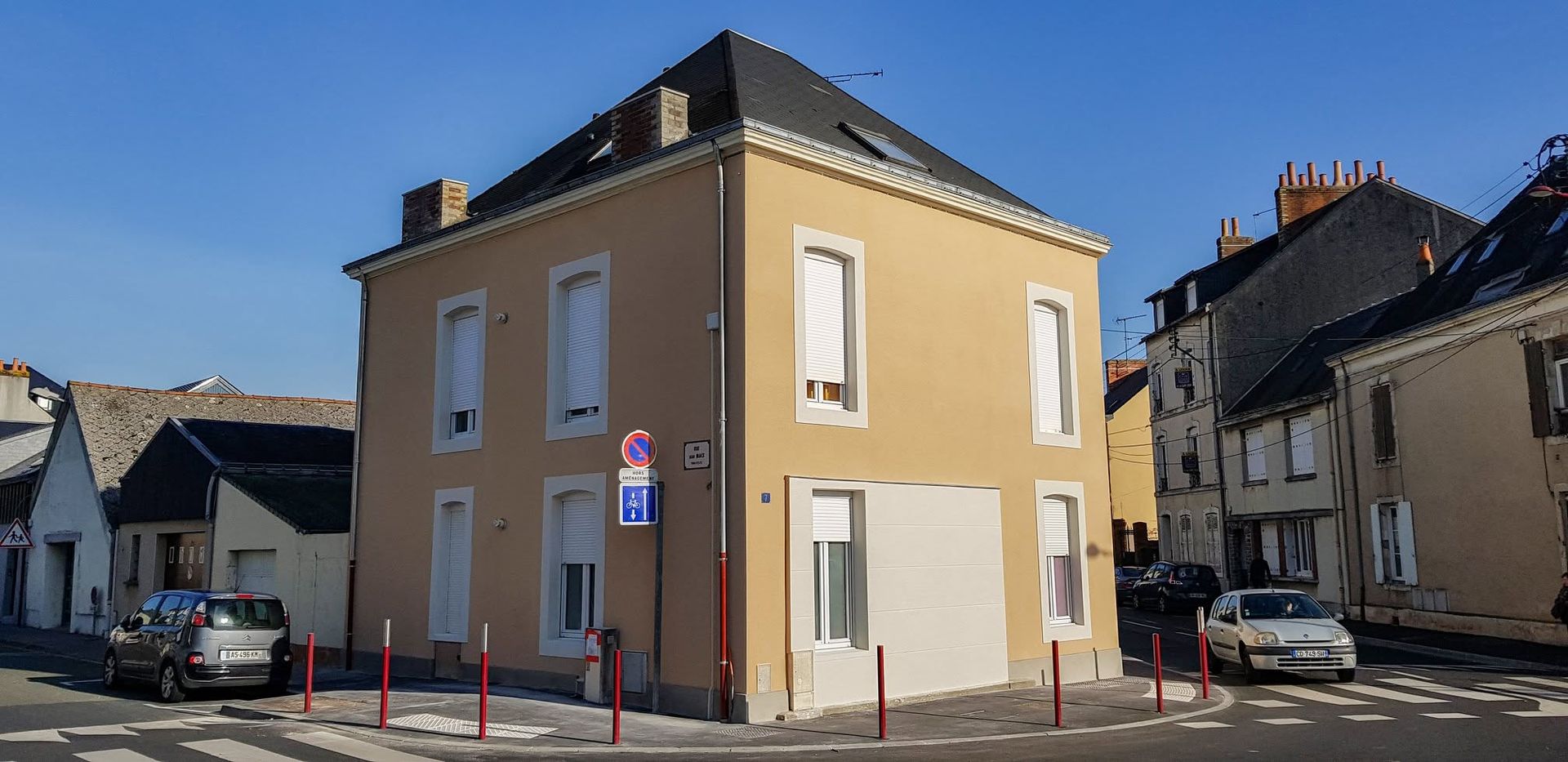 Bâtiment beige d'angle dans une rue, avec des fenêtres à encadrement blanc et un panneau de signalisation sous un ciel ensoleillé.