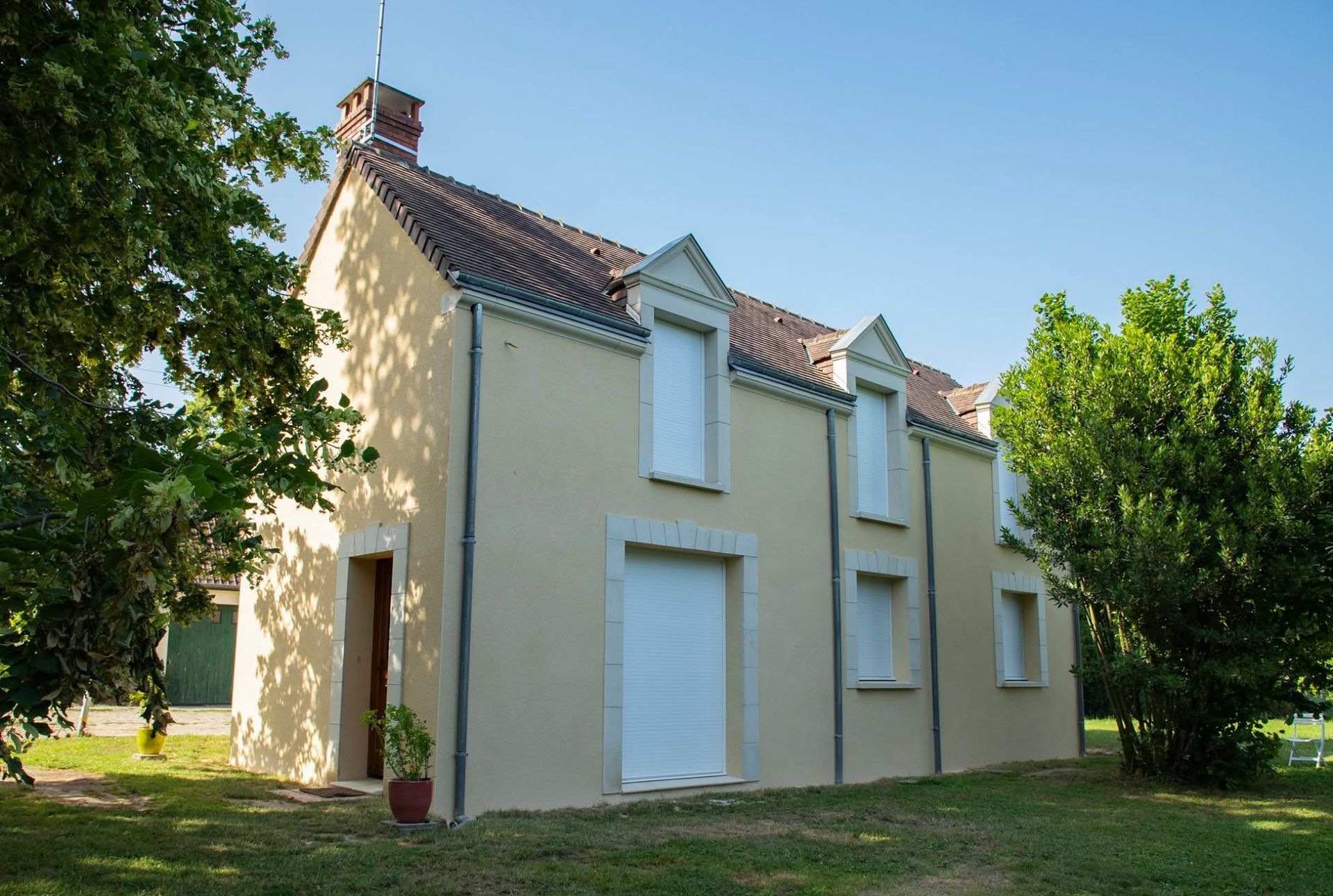 Maison beige à deux étages avec toit de tuiles rouges et volets blancs, entourée d'arbres et de pelouse.