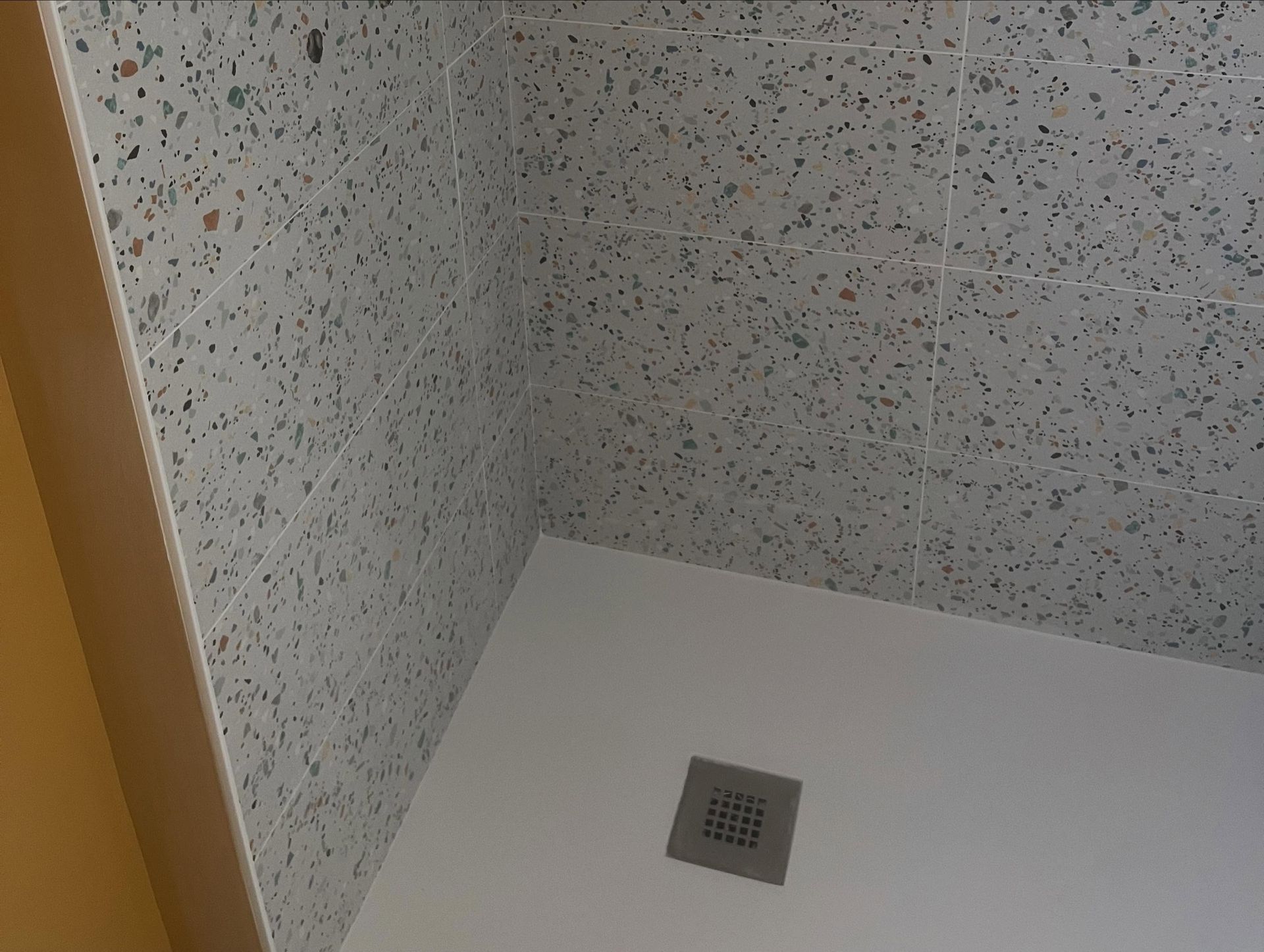 Coin d'une cabine de douche avec des parois mouchetées et une petite bonde de sol carrée.