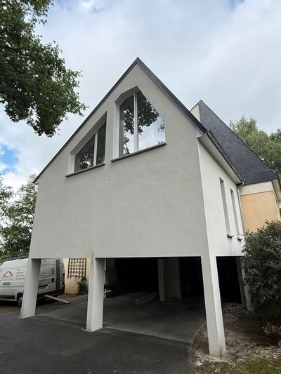 Maison moderne blanche sur pilotis, avec toit pentu et grandes fenêtres, vue de face.