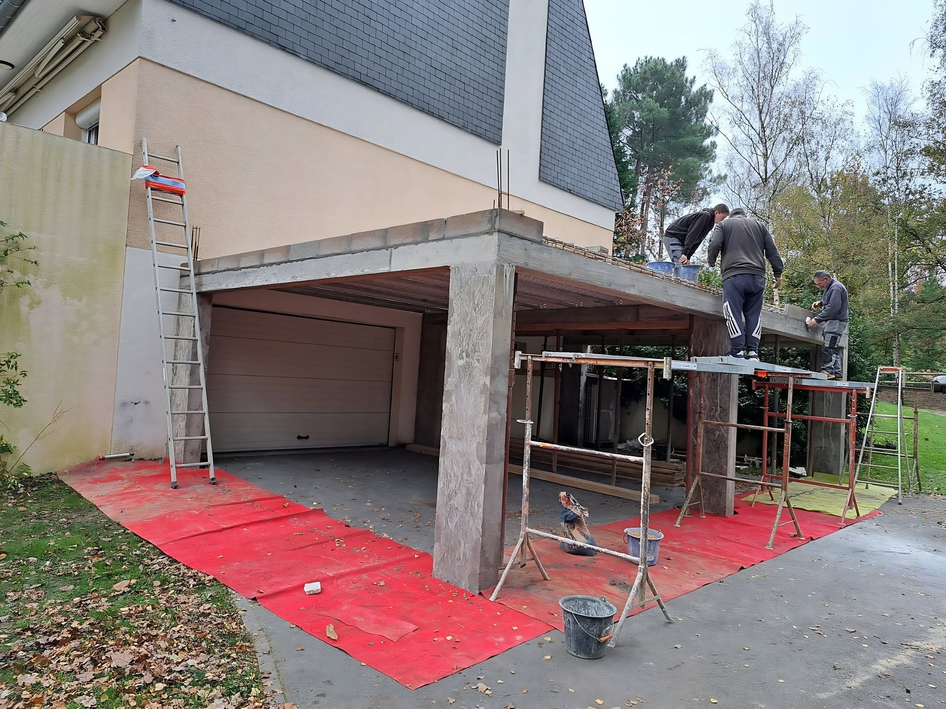 Des ouvriers travaillent sur des échafaudages pour peindre l'extérieur d'une maison et le porche d'un garage, des bâches de protection rouges étant posées au sol.