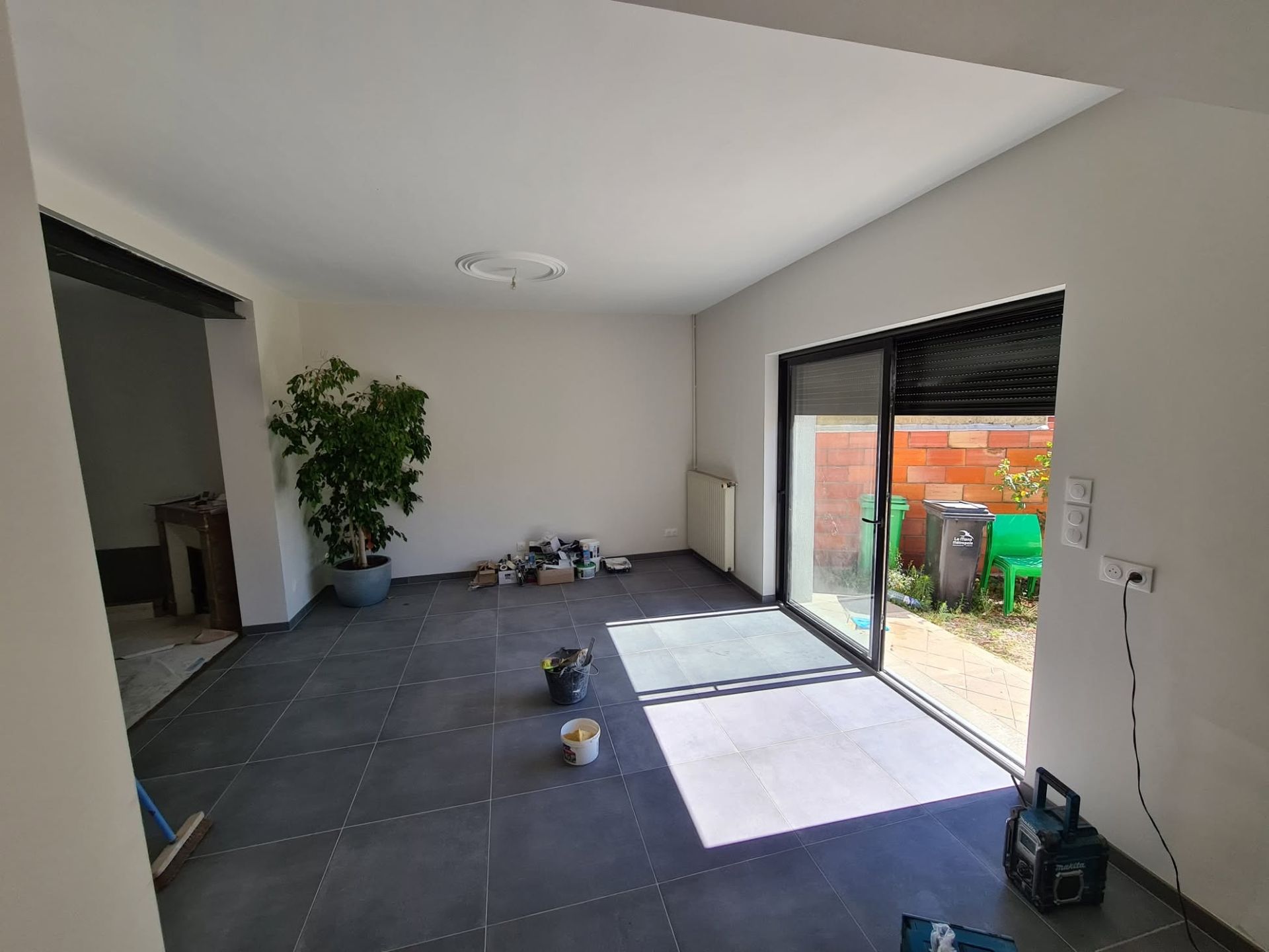 Pièce vide avec carrelage foncé au sol, plante en pot et porte coulissante ouverte donnant sur un patio avec des poubelles vertes.