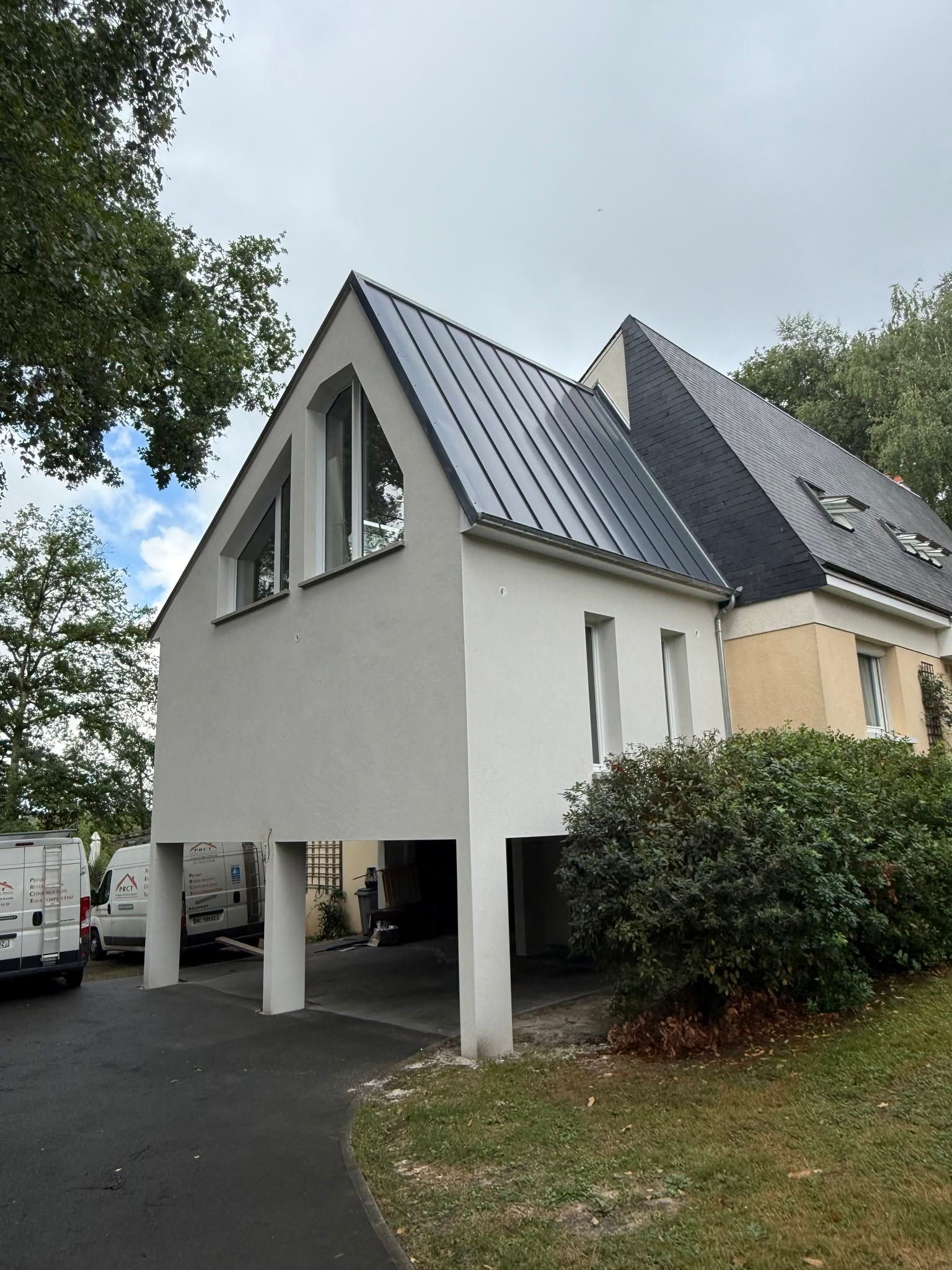 Maison blanche moderne sur pilotis avec toit foncé, allée et arbres tout autour.