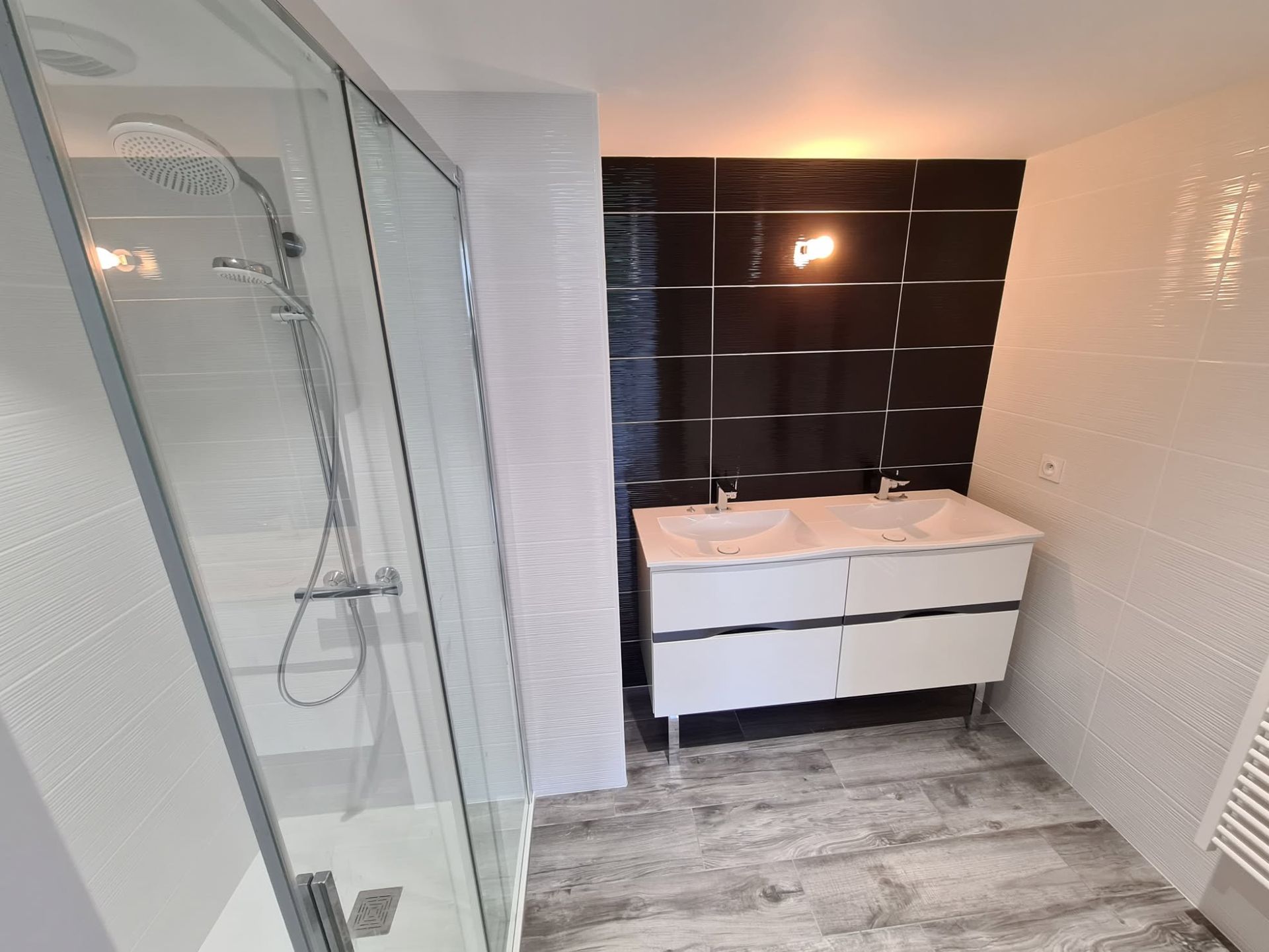 Salle de bains moderne avec douche en verre, meuble-lavabo blanc, crédence en carrelage noir et carrelage gris au sol.