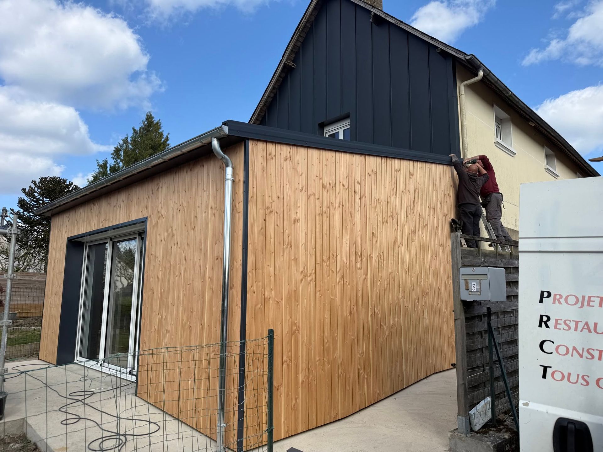 Des ouvriers installent un bardage en bois sur une extension de maison à côté d'un bâtiment blanc, sous un ciel bleu.