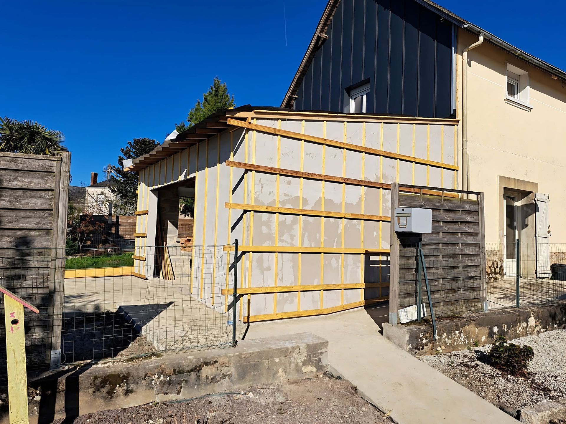 Extérieur de la maison avec échafaudages jaunes et rampe en béton sous un ciel bleu.