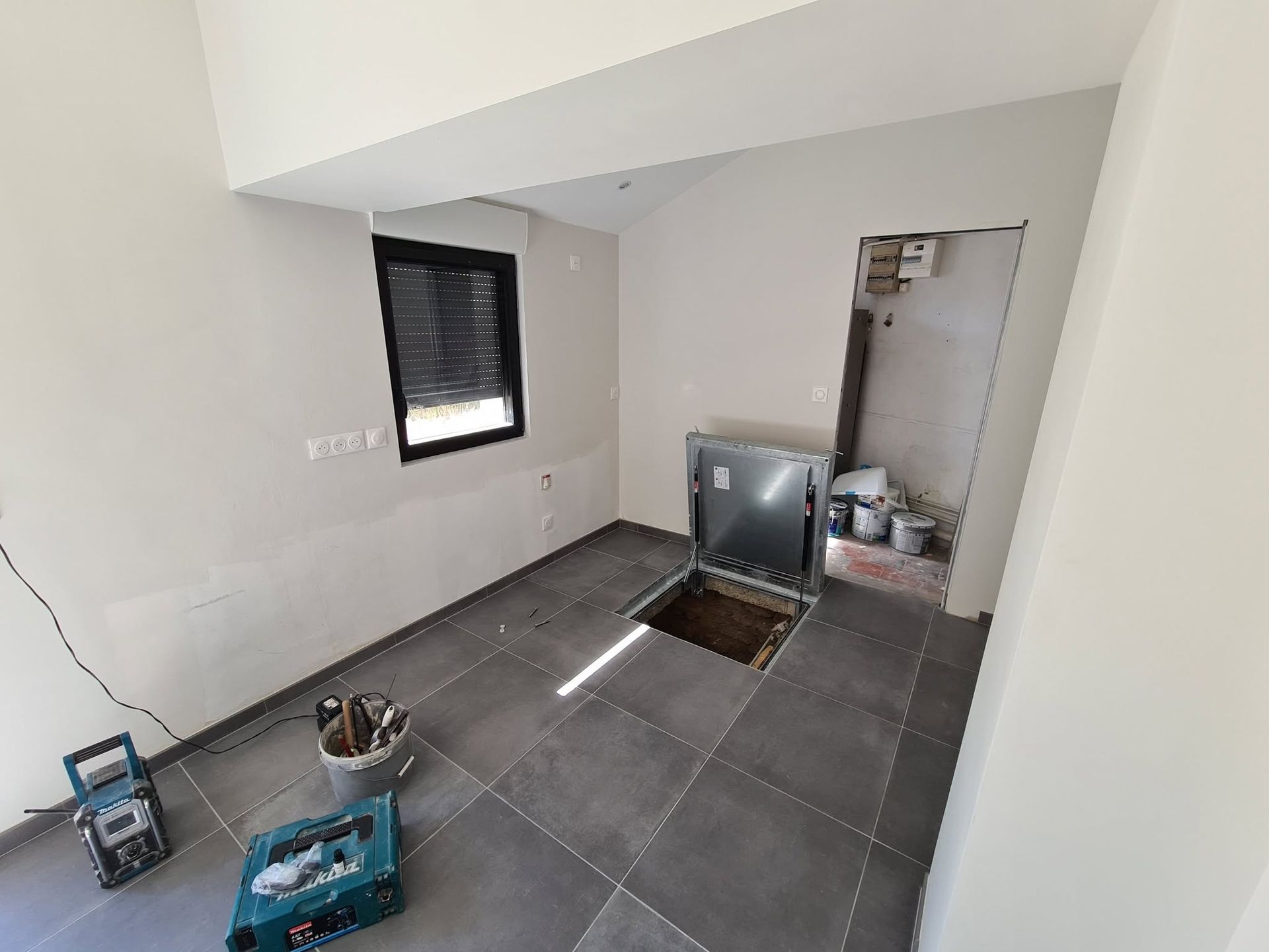 Cuisine vide en cours de rénovation, avec sol carrelé gris, évier ouvert et ustensiles éparpillés.