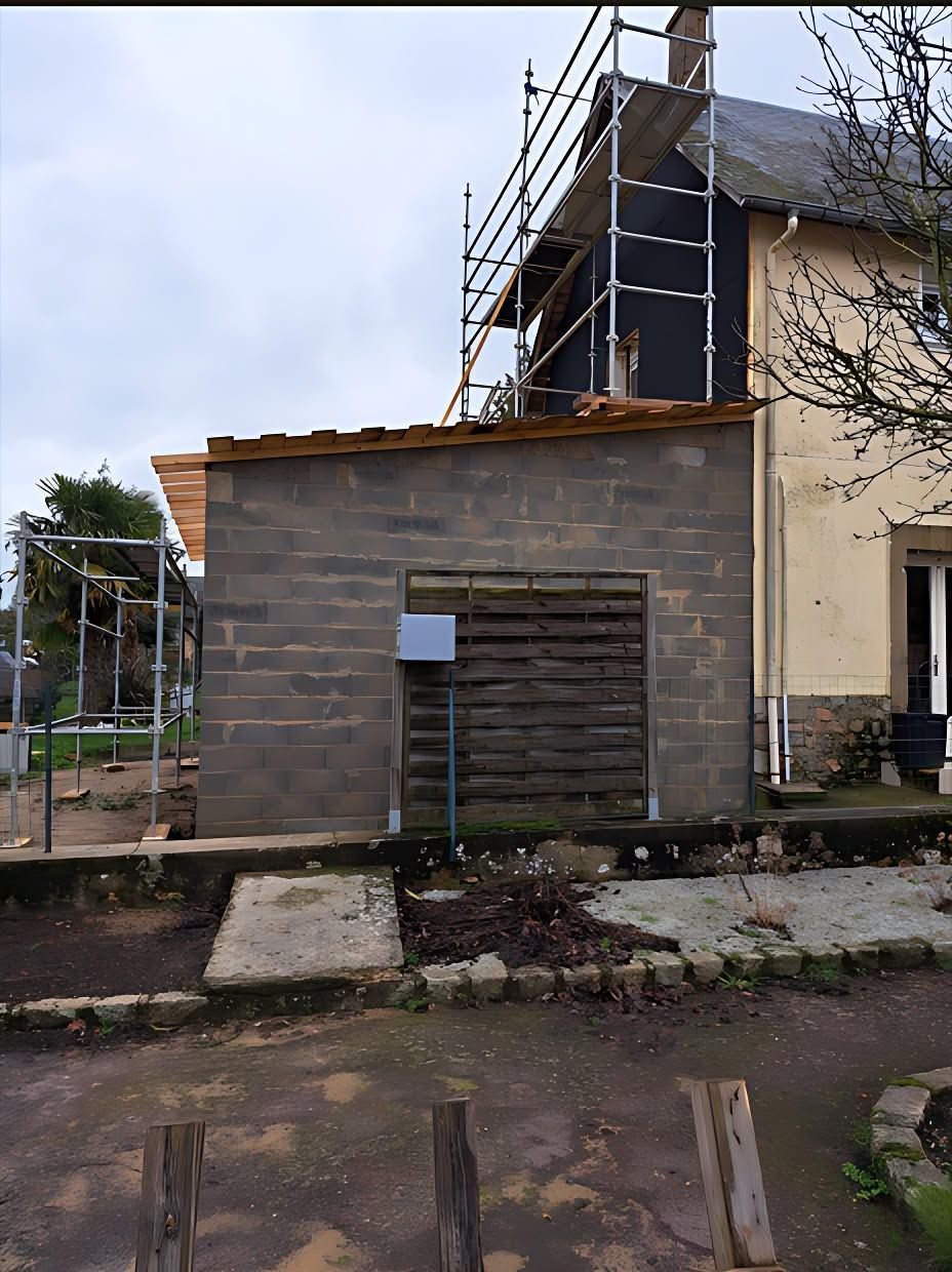 Petit hangar en parpaings à côté d'une maison en rénovation, avec des échafaudages et un chemin de terre devant.