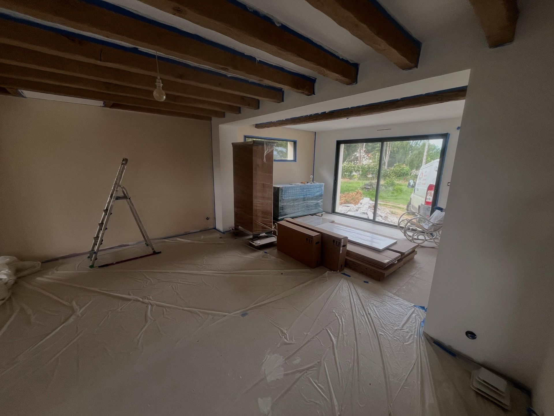 Pièce en rénovation avec poutres apparentes, échelle, sol recouvert et grande fenêtre à côté de matériaux empilés.