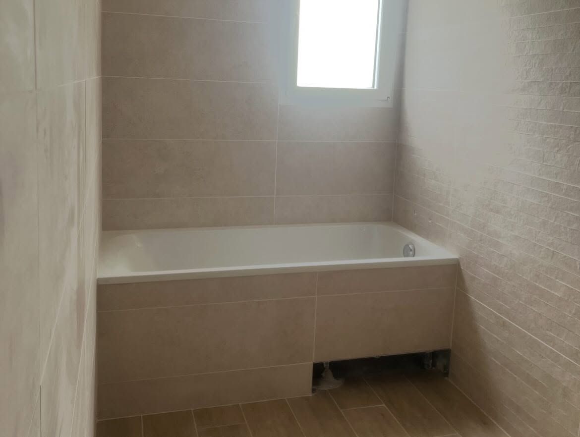Petite salle de bains carrelée de beige avec une baignoire blanche sous une fenêtre.