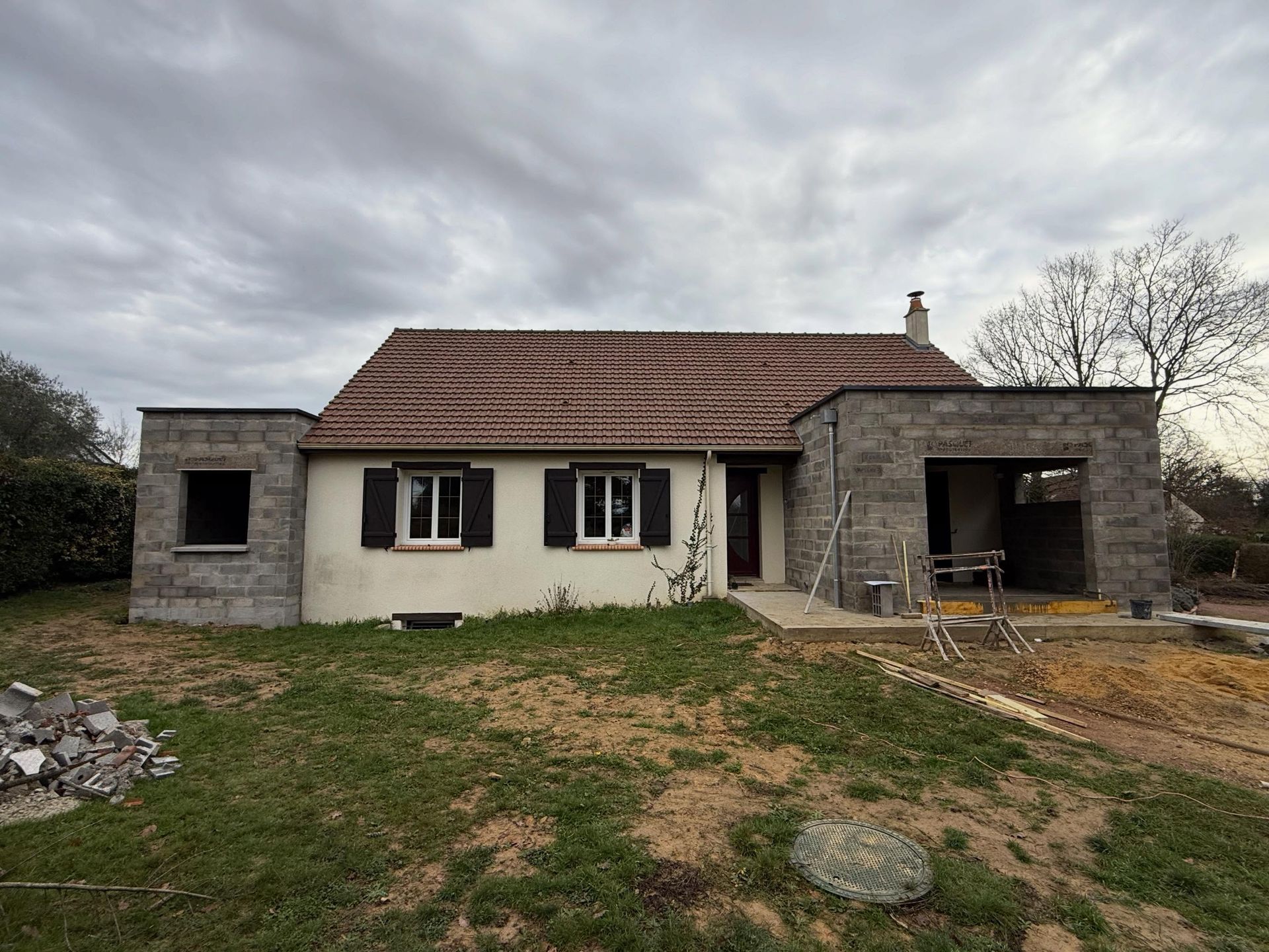 Petite maison en pierre blanche avec un toit de tuiles et un garage attenant, dans une cour herbeuse sous un ciel nuageux.