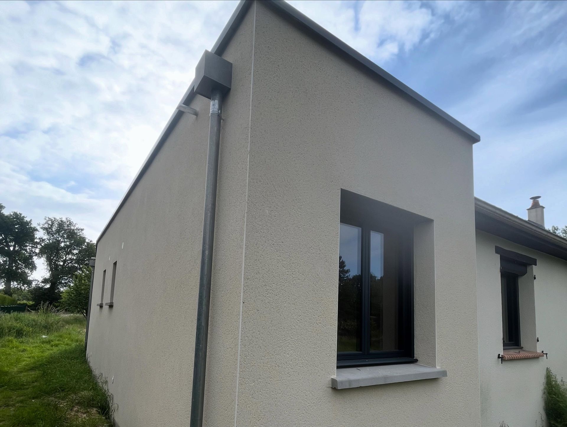 Extérieur d'une petite maison beige avec un toit en pente, des fenêtres étroites et une cour gazonnée sous un ciel nuageux.