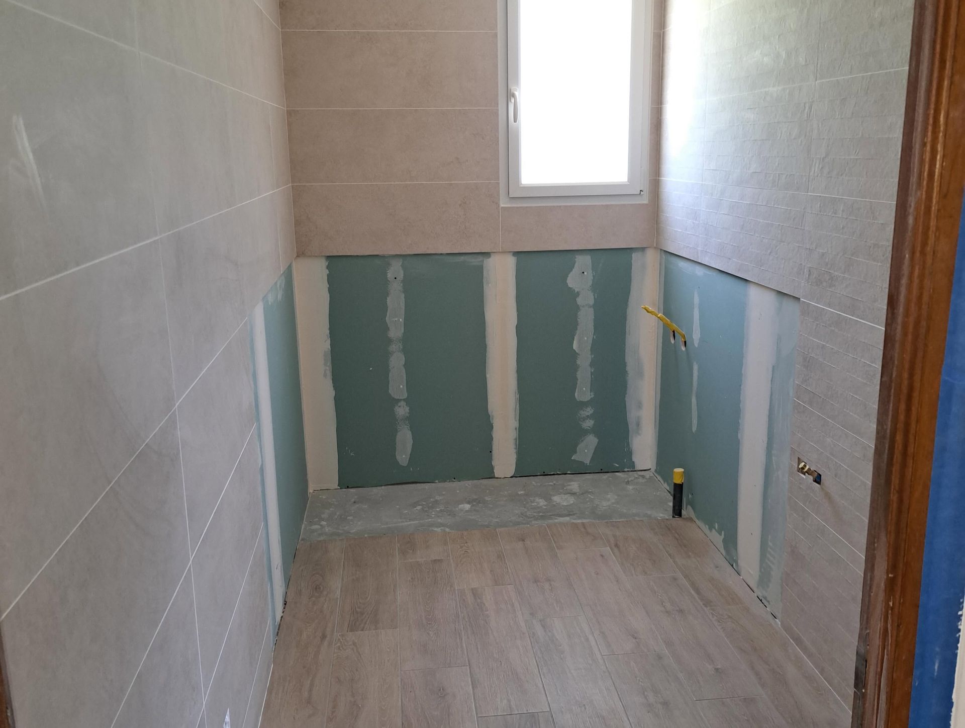 Petite salle de bains carrelée en cours de rénovation, avec des cloisons sèches vertes non finies autour d'une fenêtre et des toilettes.