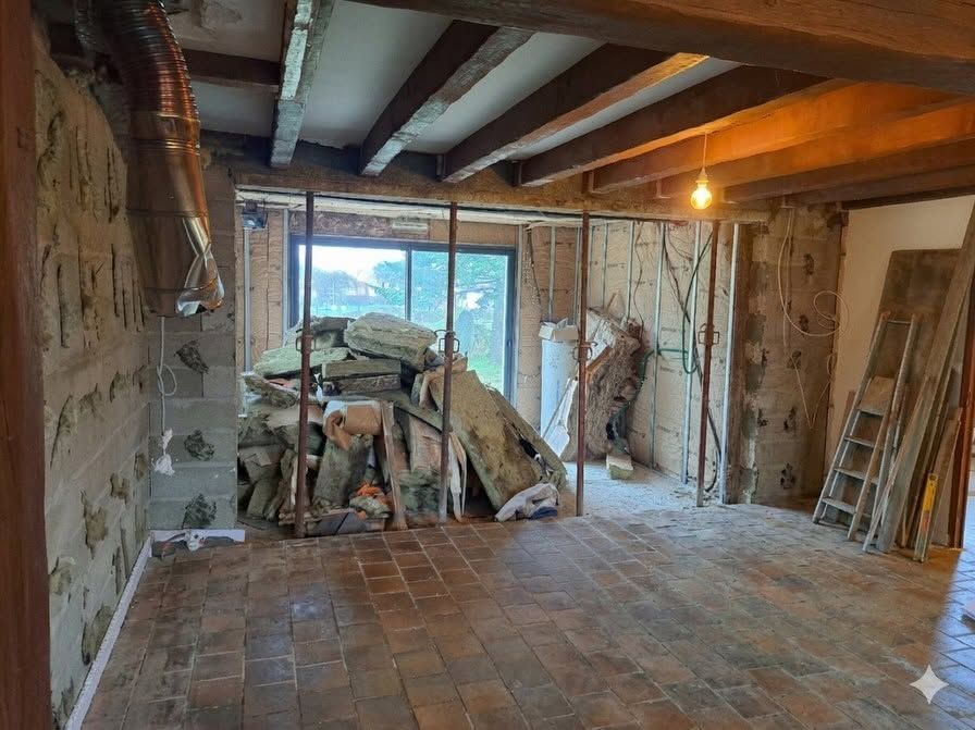 Pièce partiellement rénovée avec poutres apparentes, sol en briques et matériaux de construction près d'une fenêtre.