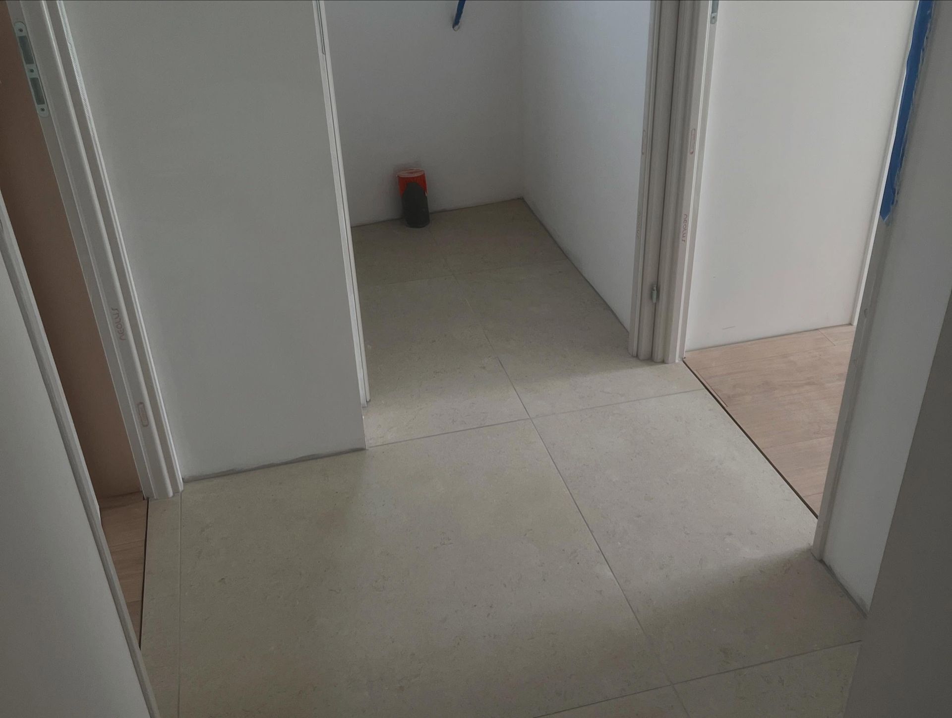 Couloir intérieur étroit aux murs blancs, au sol carrelé beige et avec un petit objet rouge et noir dans le coin du fond.
