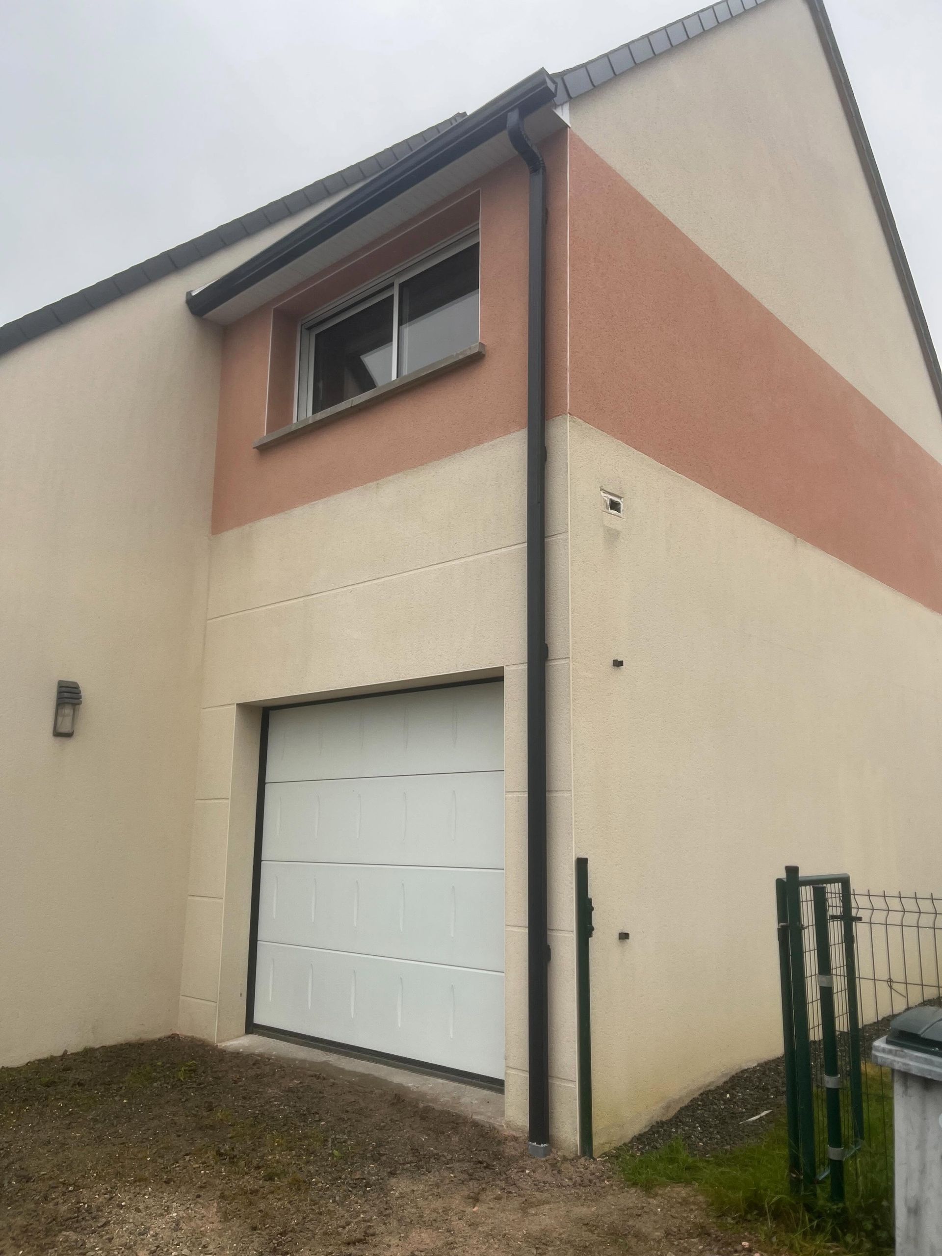 Extérieur d'une maison beige avec une porte de garage blanche et des boiseries couleur saumon sous un ciel couvert.