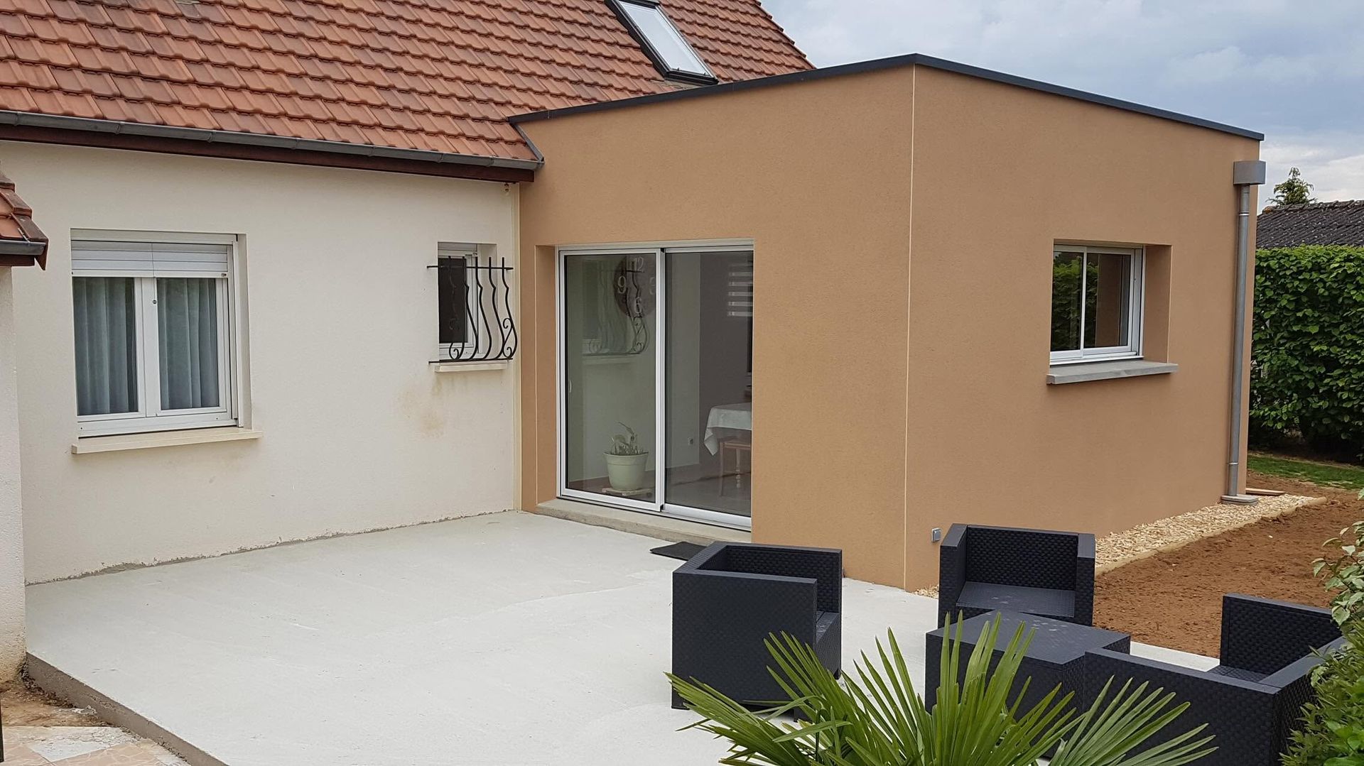 Patio de maison moderne avec murs blancs et beiges, porte-fenêtre coulissante et chaises d'extérieur sur une terrasse pavée.