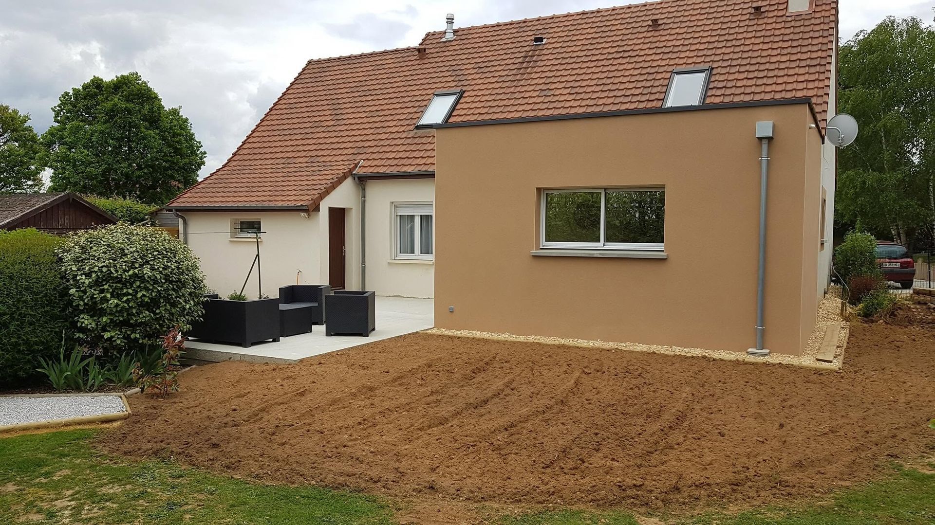 Maison avec toit de tuiles rouges et extension en stuc beige, jouxtant une cour en terre fraîchement nivelée.