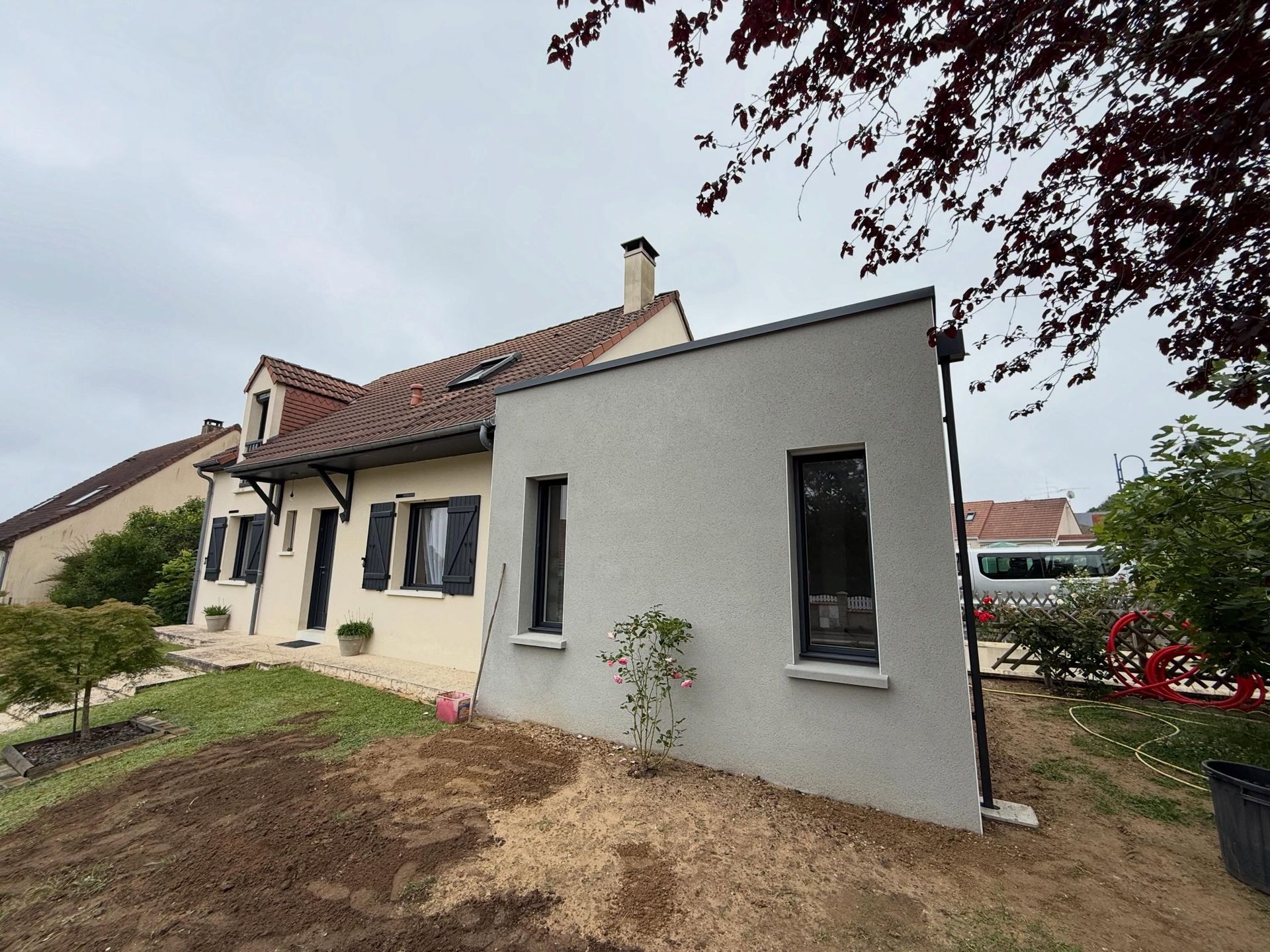 Extérieur d'une maison blanche au toit de tuiles rouges et au petit jardin sous un ciel nuageux.