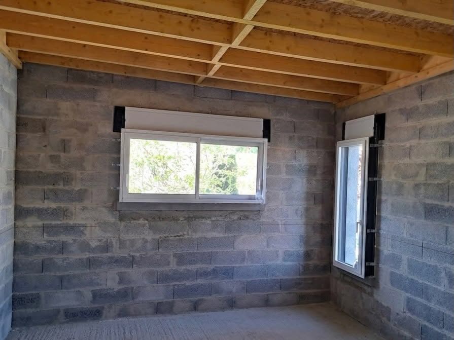 Pièce en sous-sol non aménagée avec murs en parpaings, solives de plafond en bois apparentes et deux fenêtres.