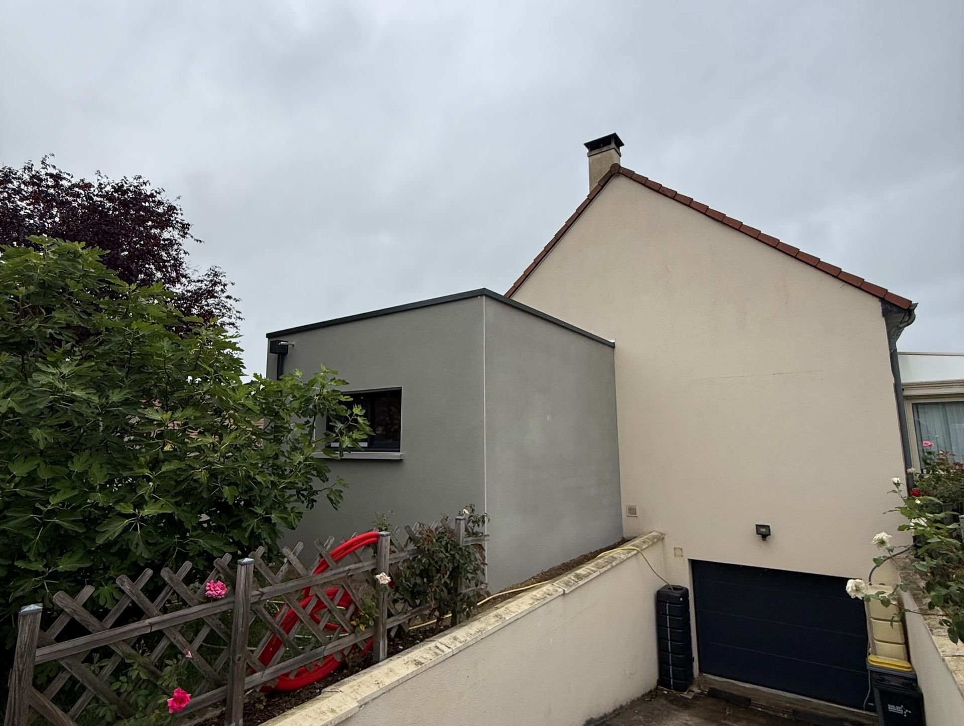 Petite maison aux murs gris et beiges, au toit en pente et au chemin de jardin sous un ciel nuageux.