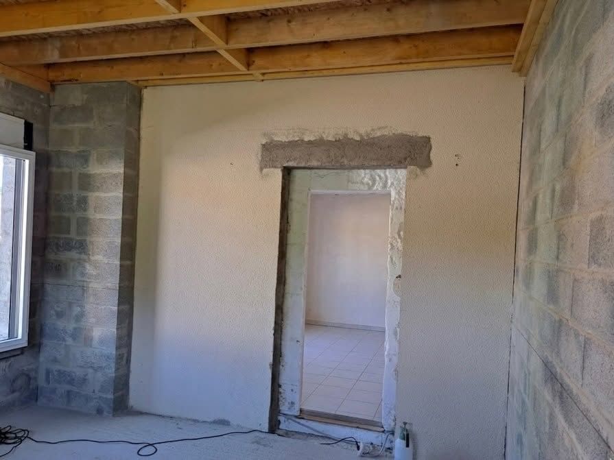 Mur intérieur partiellement fini avec une porte, poutres apparentes au plafond et côtés en parpaings.