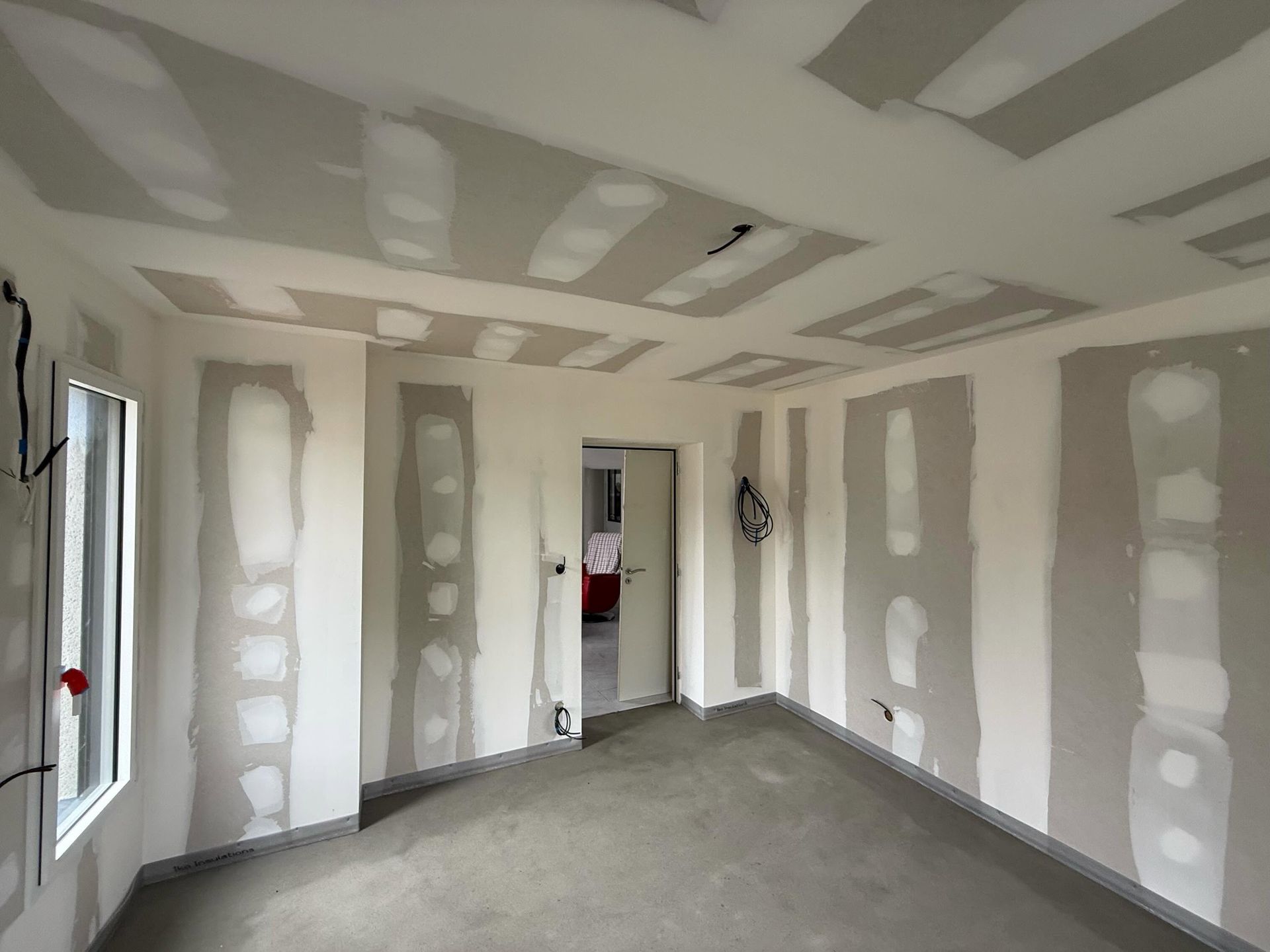 Pièce en plaques de plâtre non finies avec joints collés, sol en béton brut et une porte donnant sur une autre pièce.