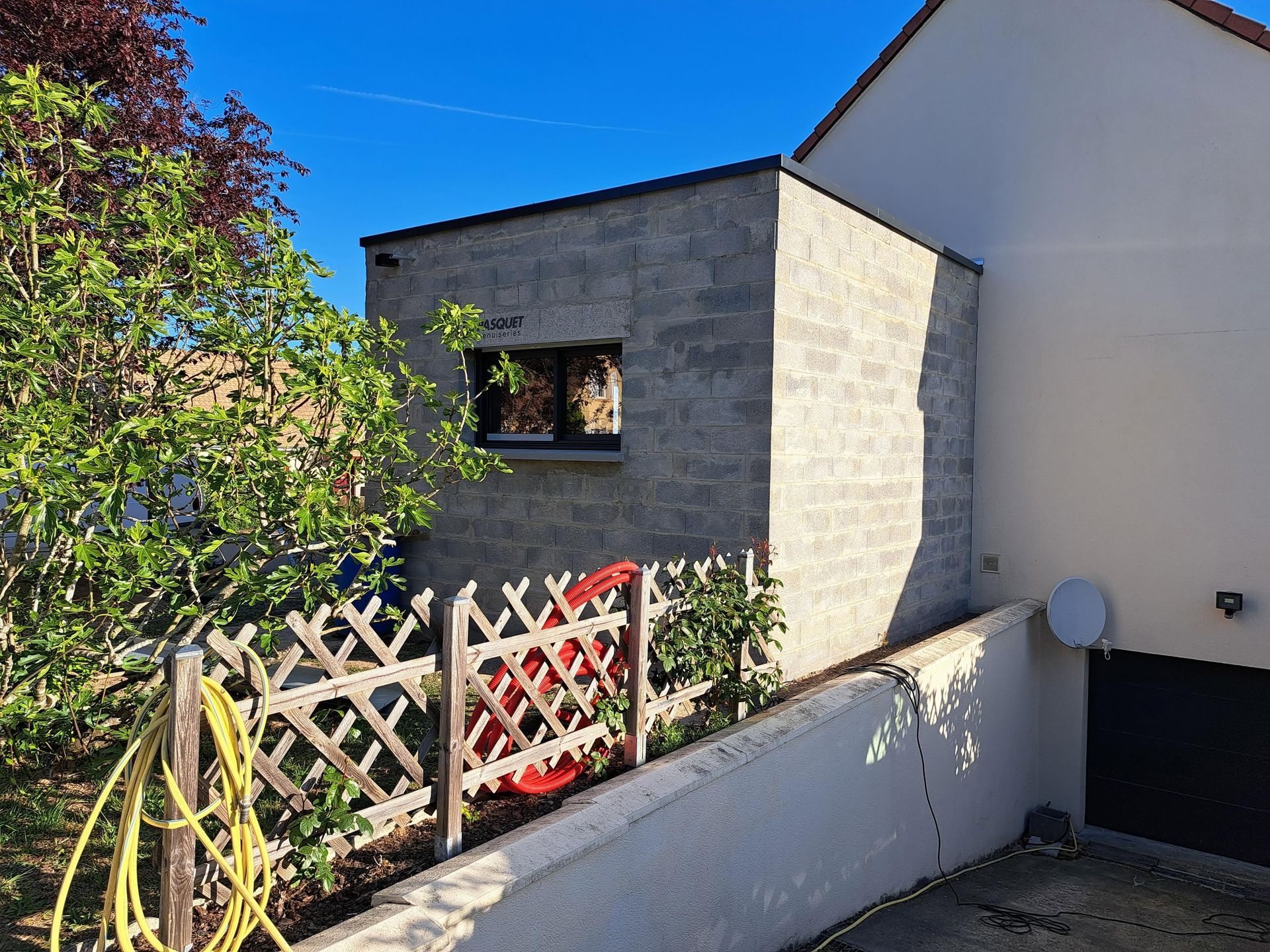 Maison en pierre avec une petite fenêtre, une clôture en bois et une allée en pente sous un ciel bleu clair.