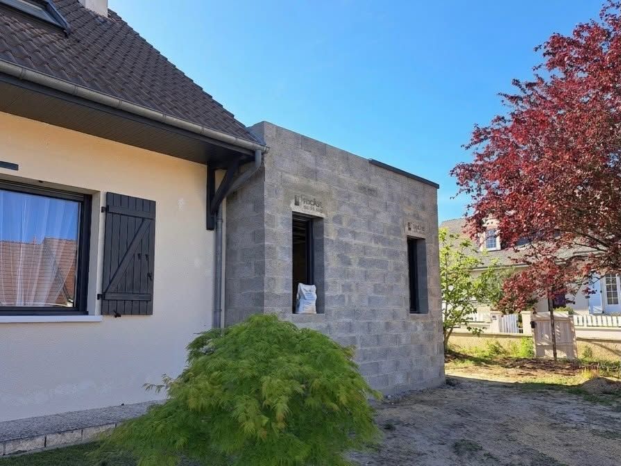 Extérieur d'une maison moderne avec murs en pierre grise, toit sombre, arbre aux feuilles rouges et ciel bleu clair.