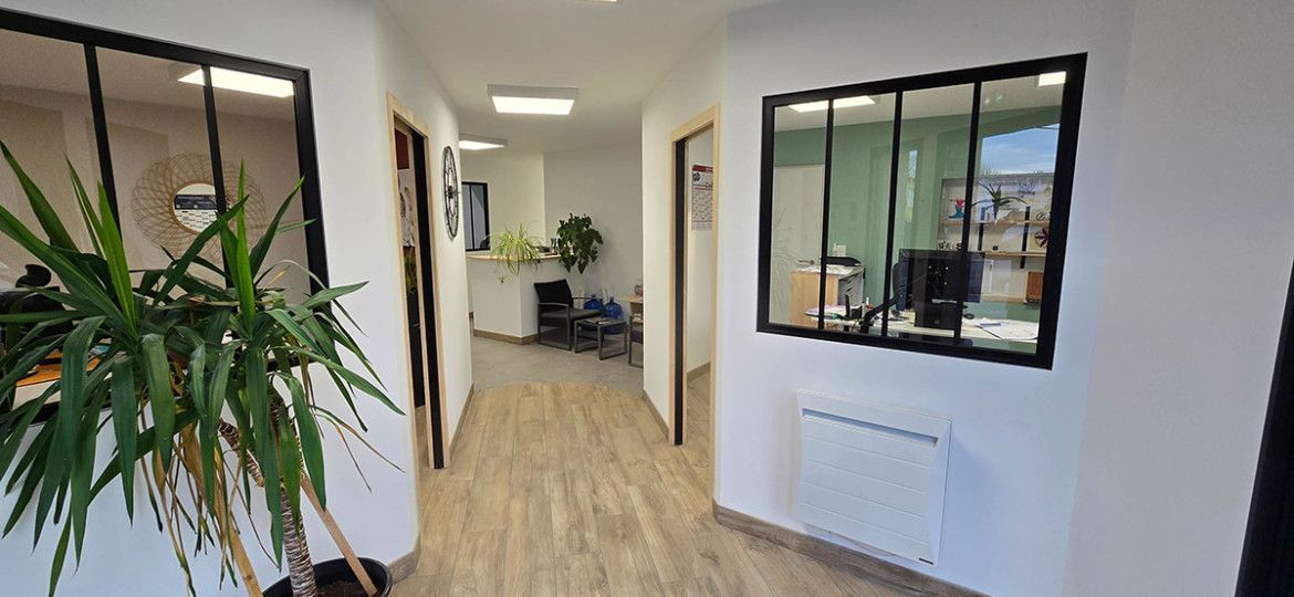 Couloir de bureau lumineux avec plante en pot, parquet, murs blancs et salle de réunion vitrée.