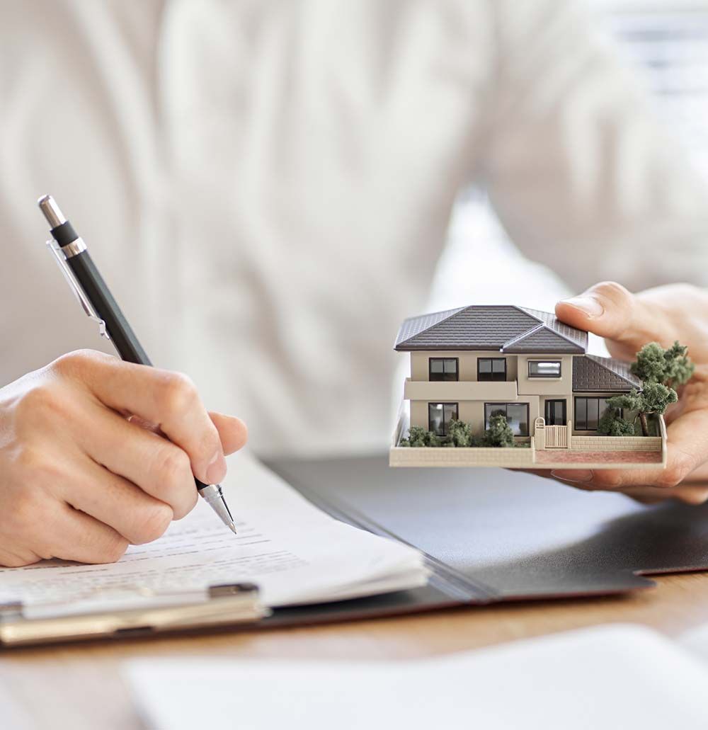 Persona escribiendo en papel, sosteniendo una maqueta de casa; probablemente una transacción inmobiliaria.