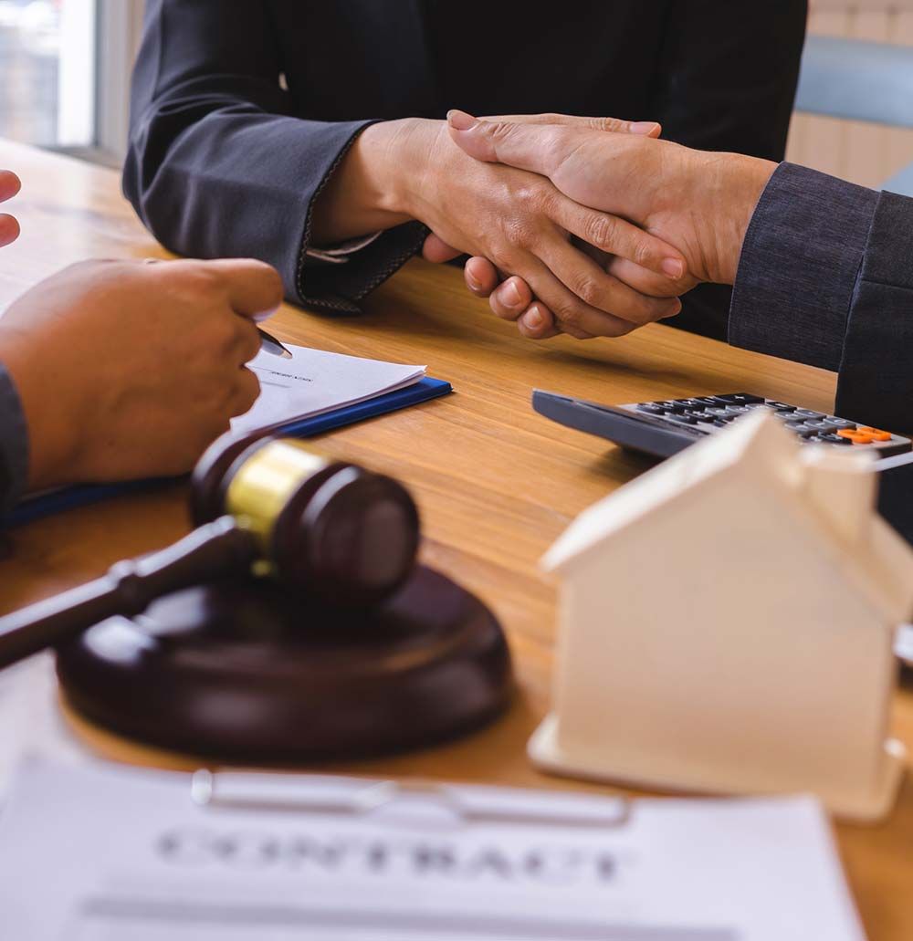 Gente dándose la mano para firmar un contrato, con un mazo y una casa de juguete sobre la mesa.