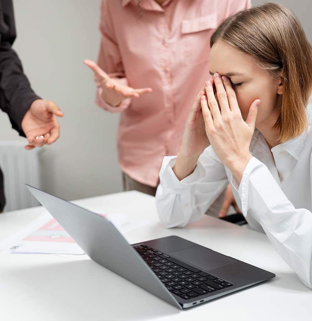 Mujer estresada en el escritorio, con las manos en la cara y dos personas gesticulando detrás de ella.