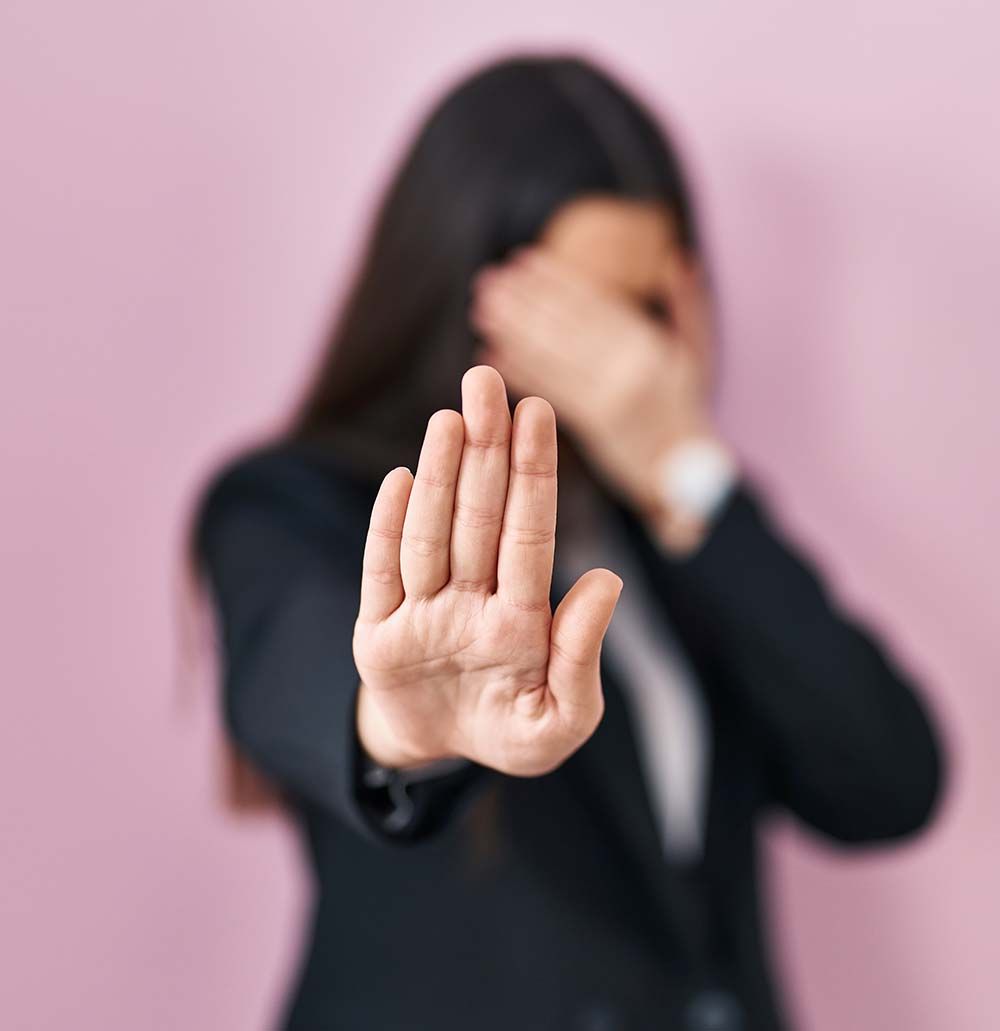 Mujer con traje, tapándose la cara con una mano y extendiendo la otra, con la palma hacia adelante.