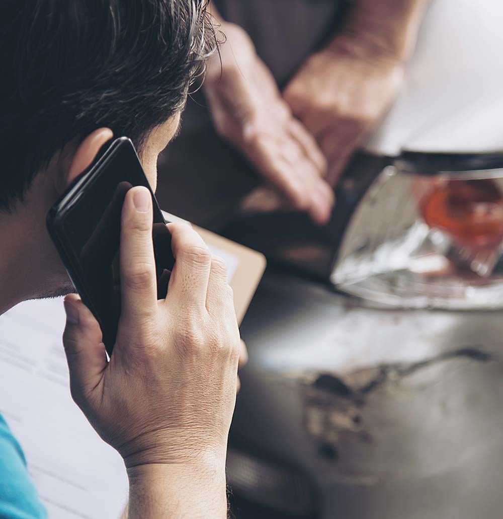 Un hombre al teléfono mira un coche averiado. Hay otra persona cerca.