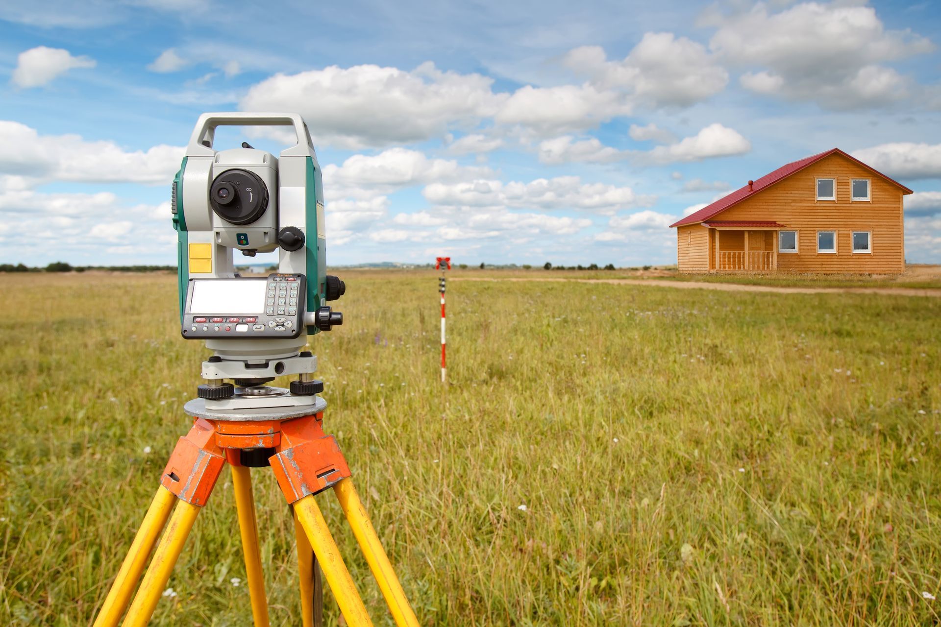 Equipo de topografía sobre un trípode en un campo, casa de madera al fondo, cielo azul con nubes.
