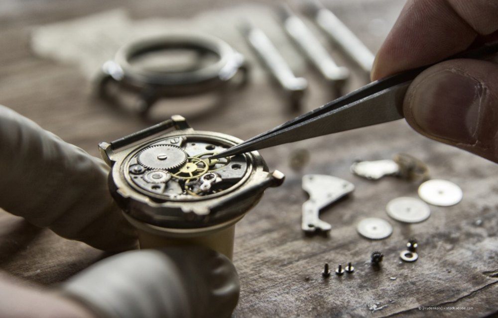 Ein Uhrmacher justiert mit einer Pinzette vorsichtig die filigranen Zahnräder und Teile einer zerlegten mechanischen Armbanduhr.