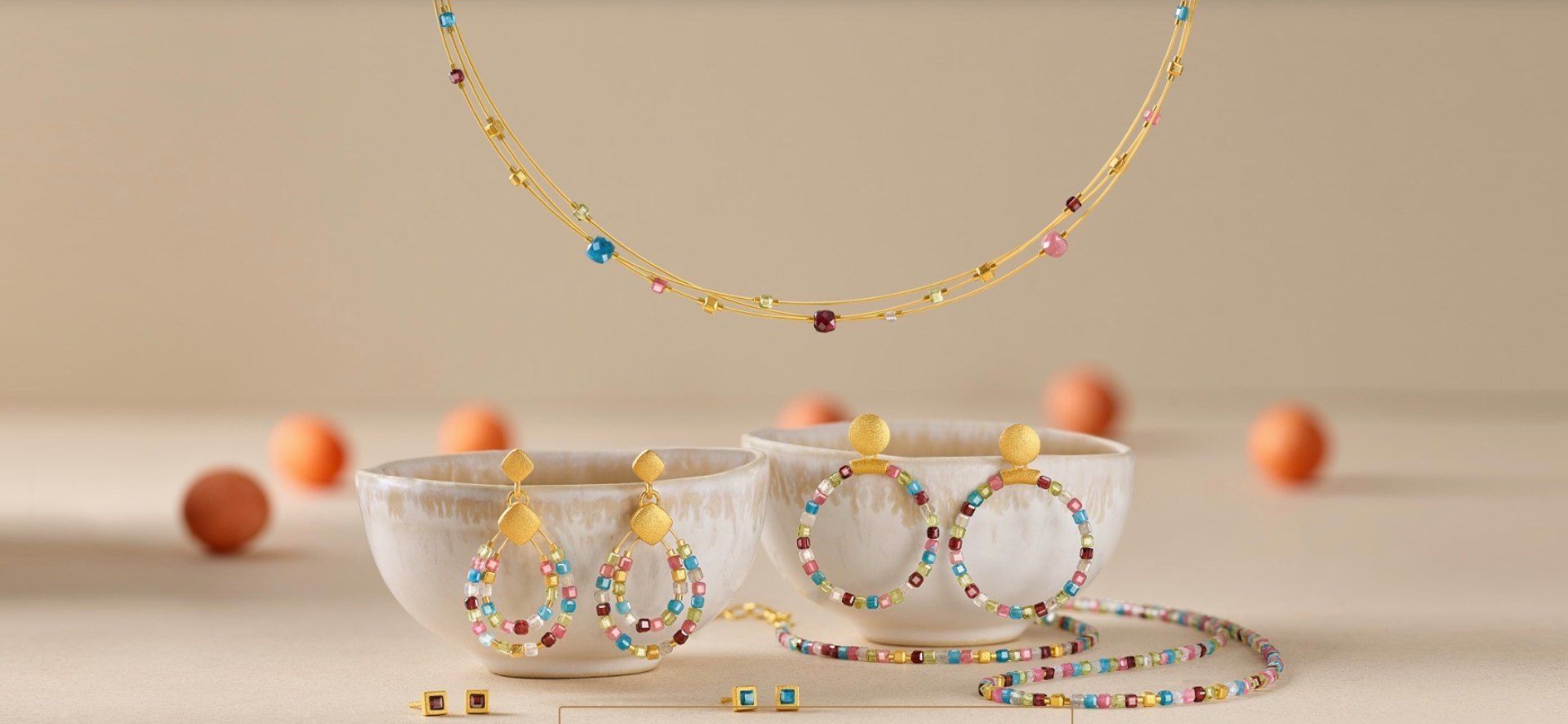 Eine goldene Halskette mit bunten Edelsteinen hängt über zwei kleinen weißen Schalen, in denen passende Creolen auf einer beigen Oberfläche liegen.