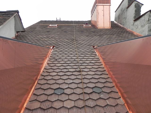 Ein gelbes Haus mit rotem Ziegeldach, blauen Fensterläden, einem kleinen Balkon mit Markise und einer hohen Konifere in der Nähe.