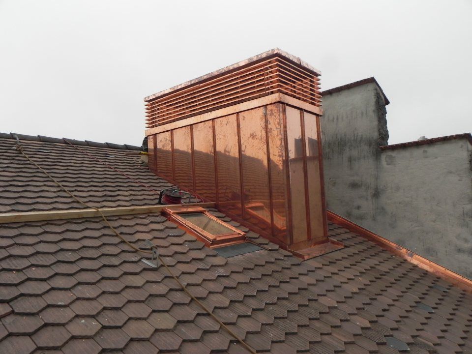 Ein mit Kupfer verkleideter Schornstein steht auf einem braunen Ziegeldach neben einem Oberlicht und einer grauen Steinmauer.