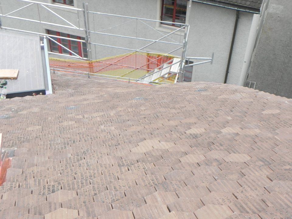 Nahaufnahme eines mit Schindeln gedeckten Daches in neutralen Farbtönen, Blickrichtung Gerüst und Mauer im Hintergrund.