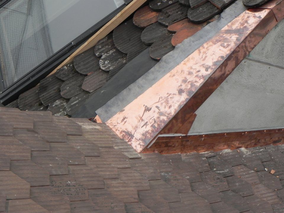 Ein Kupferblechstreifen verbindet eine mit Schindeln gedeckte Dachschräge mit einer senkrechten Wand und einem Abschnitt mit geschwungenen Dachziegeln.