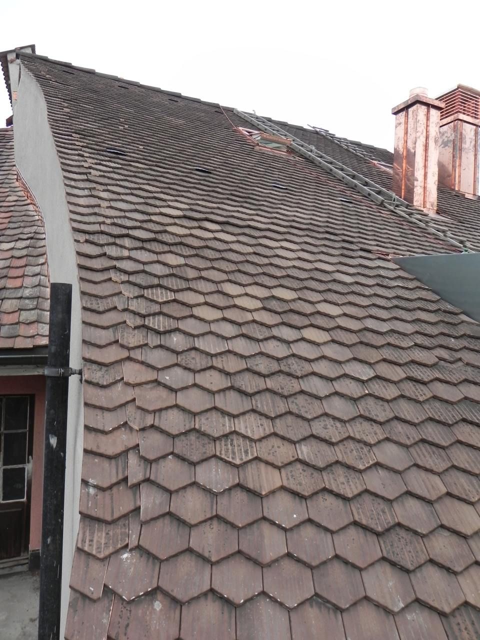 Nahaufnahme eines verwitterten, schrägen Daches, das mit sechseckigen braunen Ziegeln gedeckt ist, mit einem Schornstein und der Hausverkleidung.