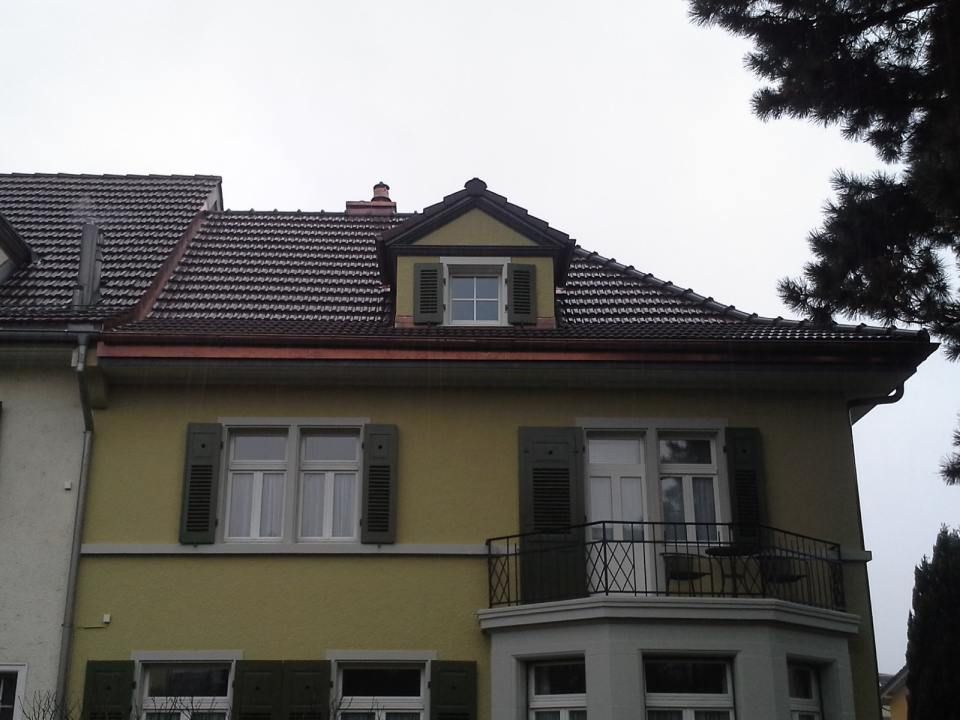 Ein senfgelbes zweistöckiges Haus mit braunen Dachziegeln, einem Dachgaubenfenster, grünen Fensterläden und einem kleinen Balkon.