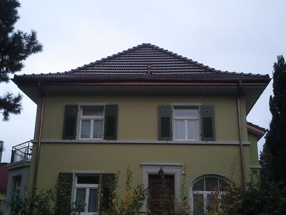 Ein zweistöckiges, gelb getünchtes Haus mit braunem Walmdach, dunklen Fensterläden und einem teilweise verdeckten Eingang.